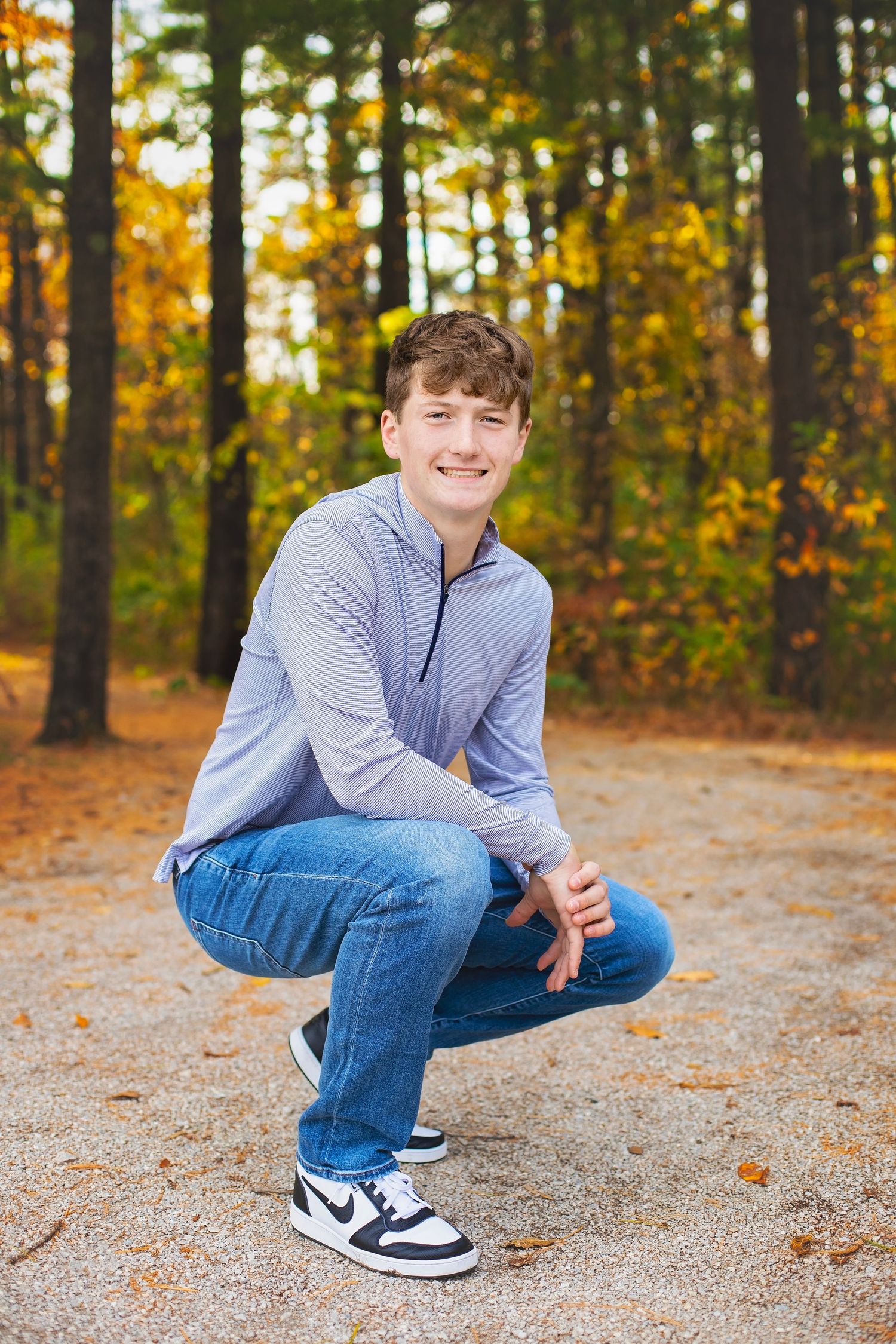 Smiling teen boy crouching in Nike sneakers and jeans outdoors among autumn foliage for senior photos.