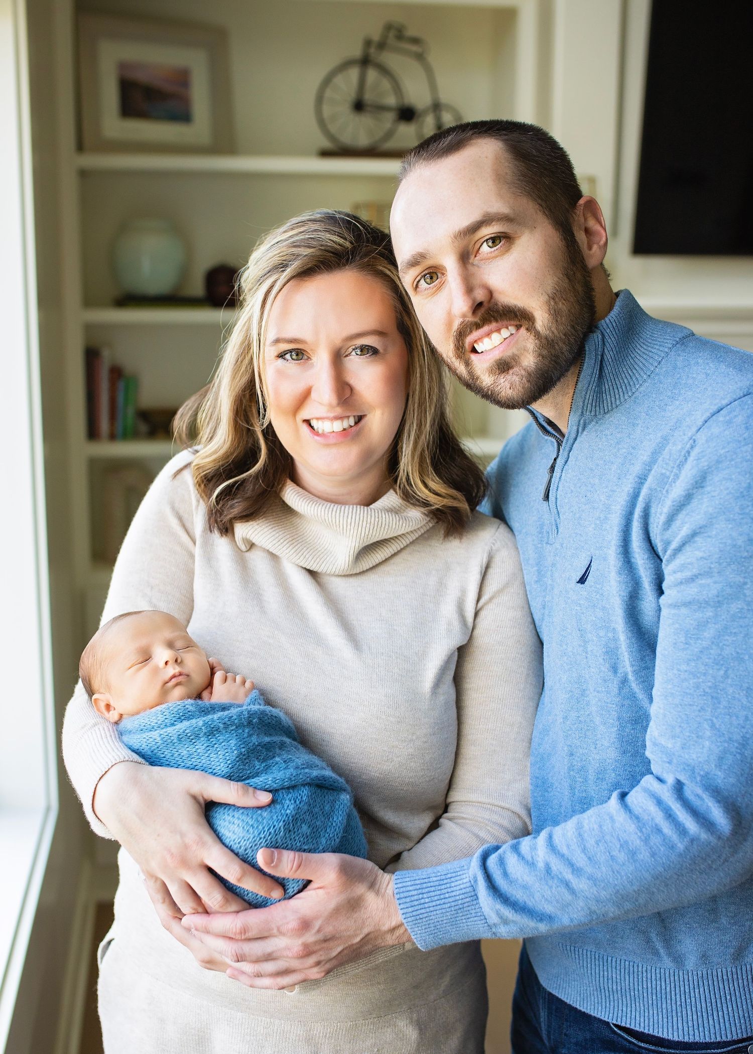 Smiling couple posing indoors holding their newborn baby wrapped in a blue swaddle blanket.