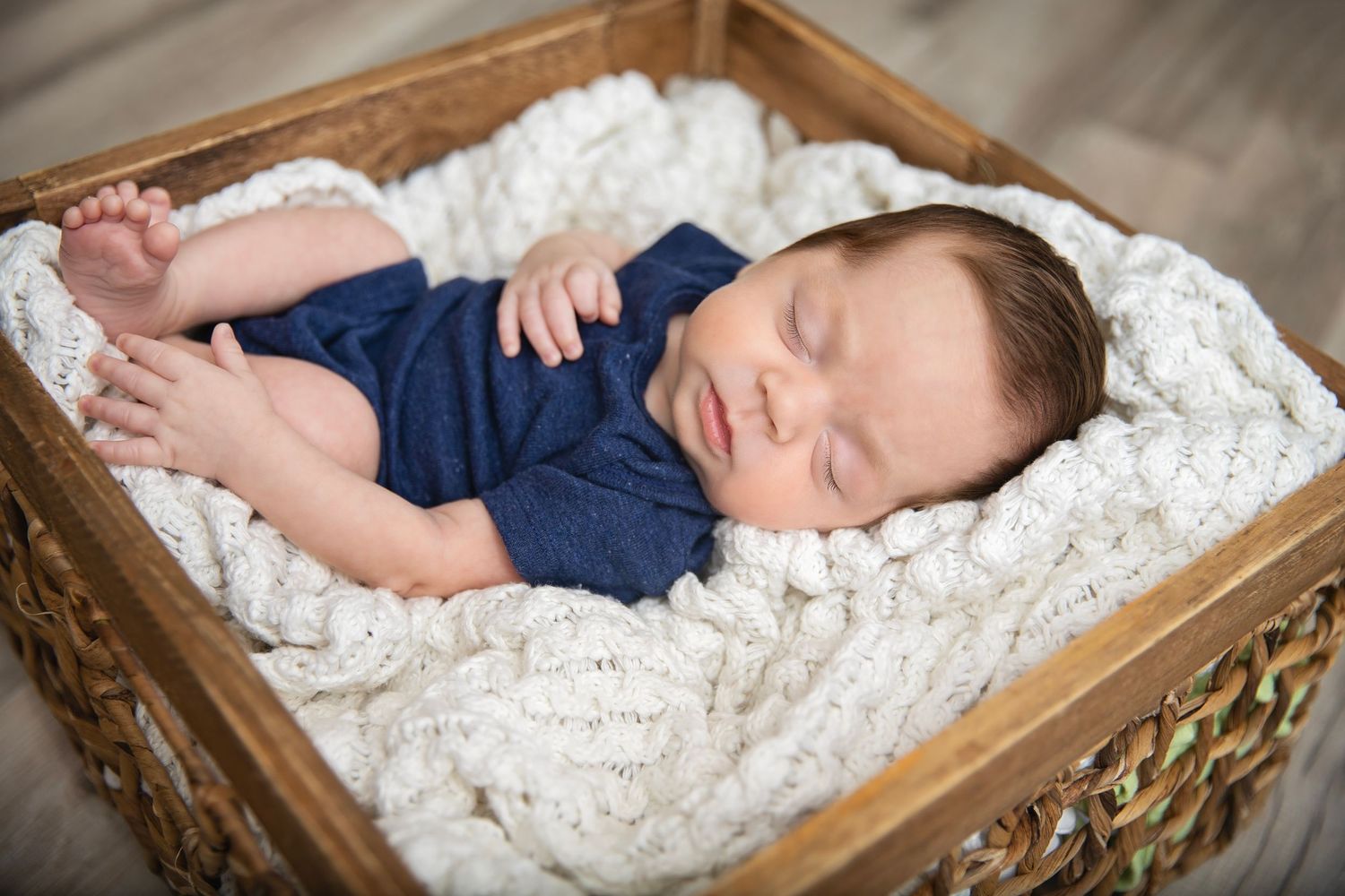 Newborn baby sleeping in a wooden basket lined with white chunky knit blanket in blue wrap.