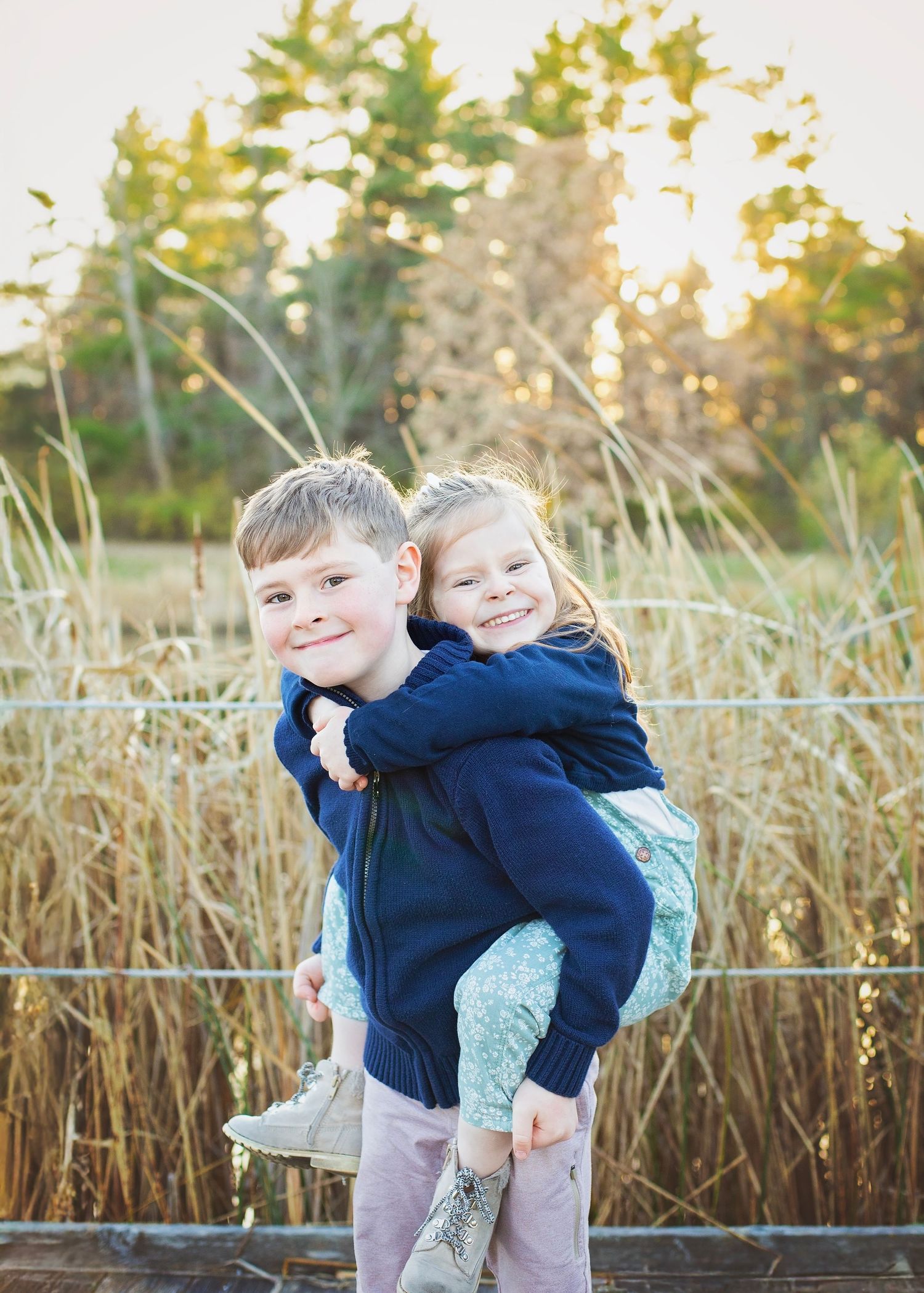 Young boy giving smiling little girl a piggyback ride outdoors near tall golden grass at sunset.