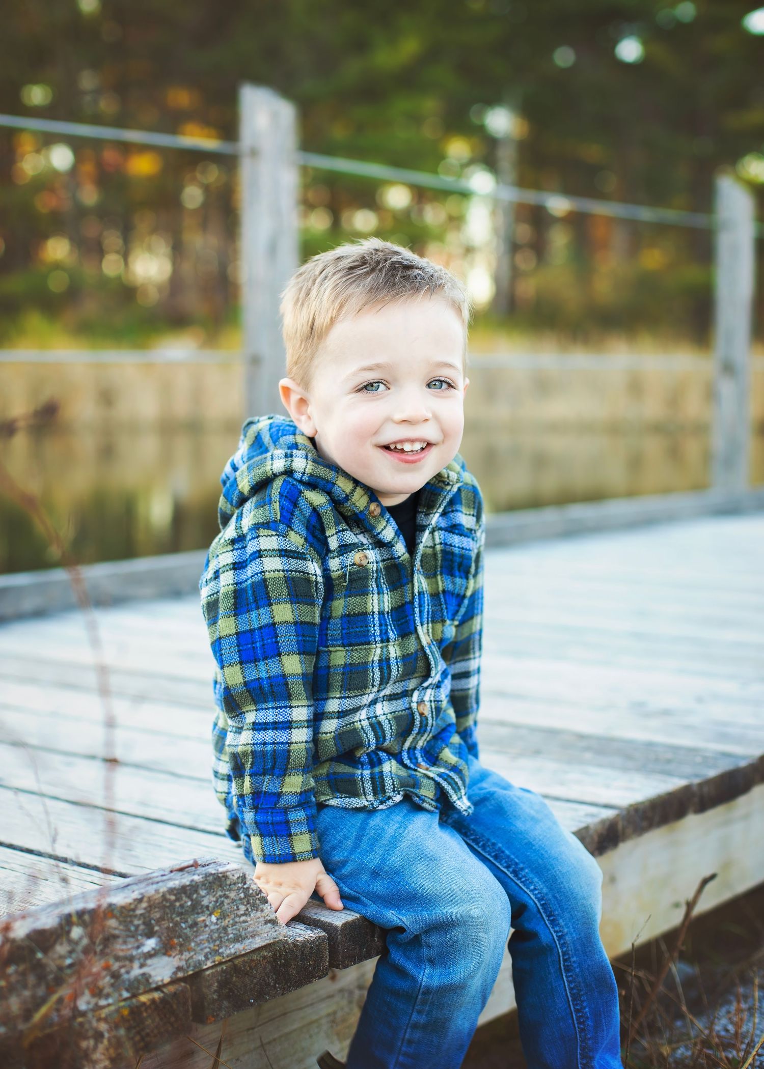 Cheerful young boy in plaid shirt sitting on wooden boardwalk edge smiling outdoors in fall setting.