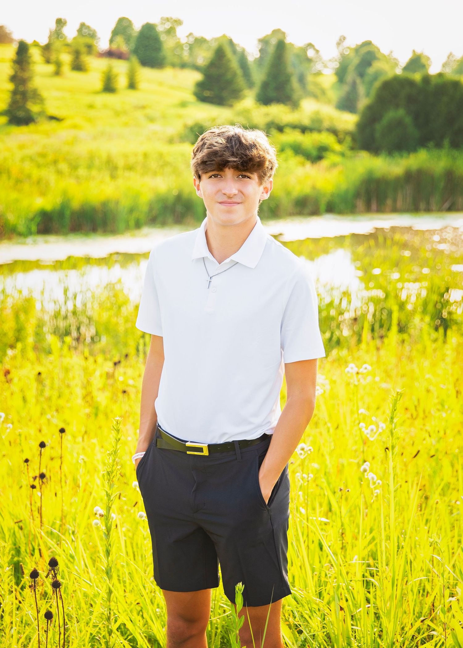 Teen boy in white polo shirt and dark shorts standing in a vibrant green meadow with a pond in the background.