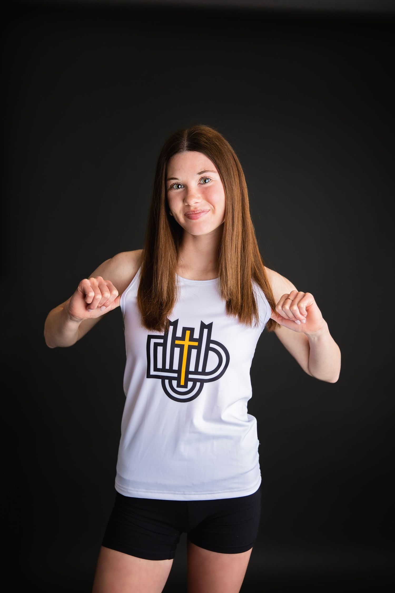 Smiling young female athlete in white tank top with cross logo pointing downward against dark background.
