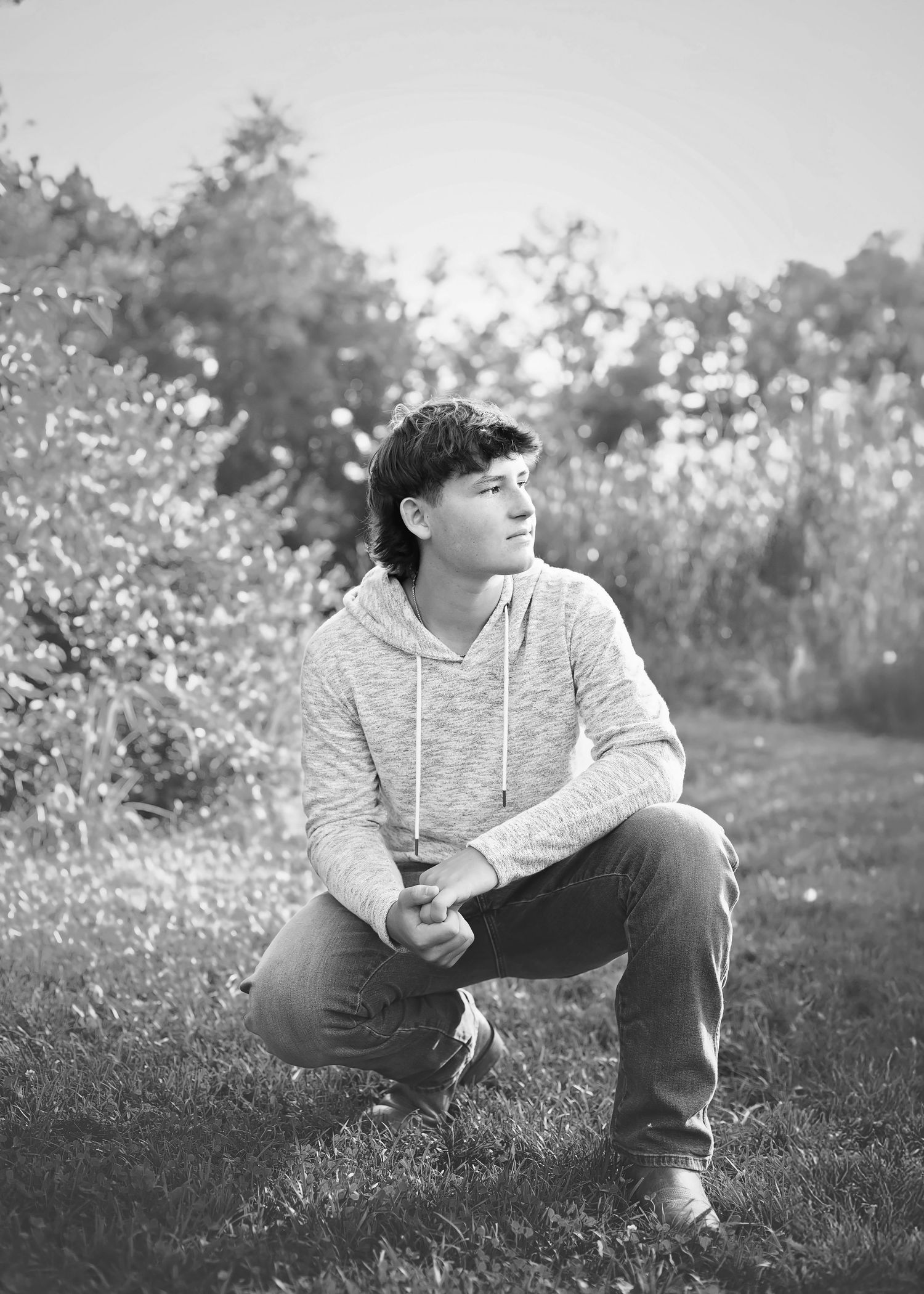 Black and white portrait of a teenage boy crouching in a field wearing a hoodie and jeans.