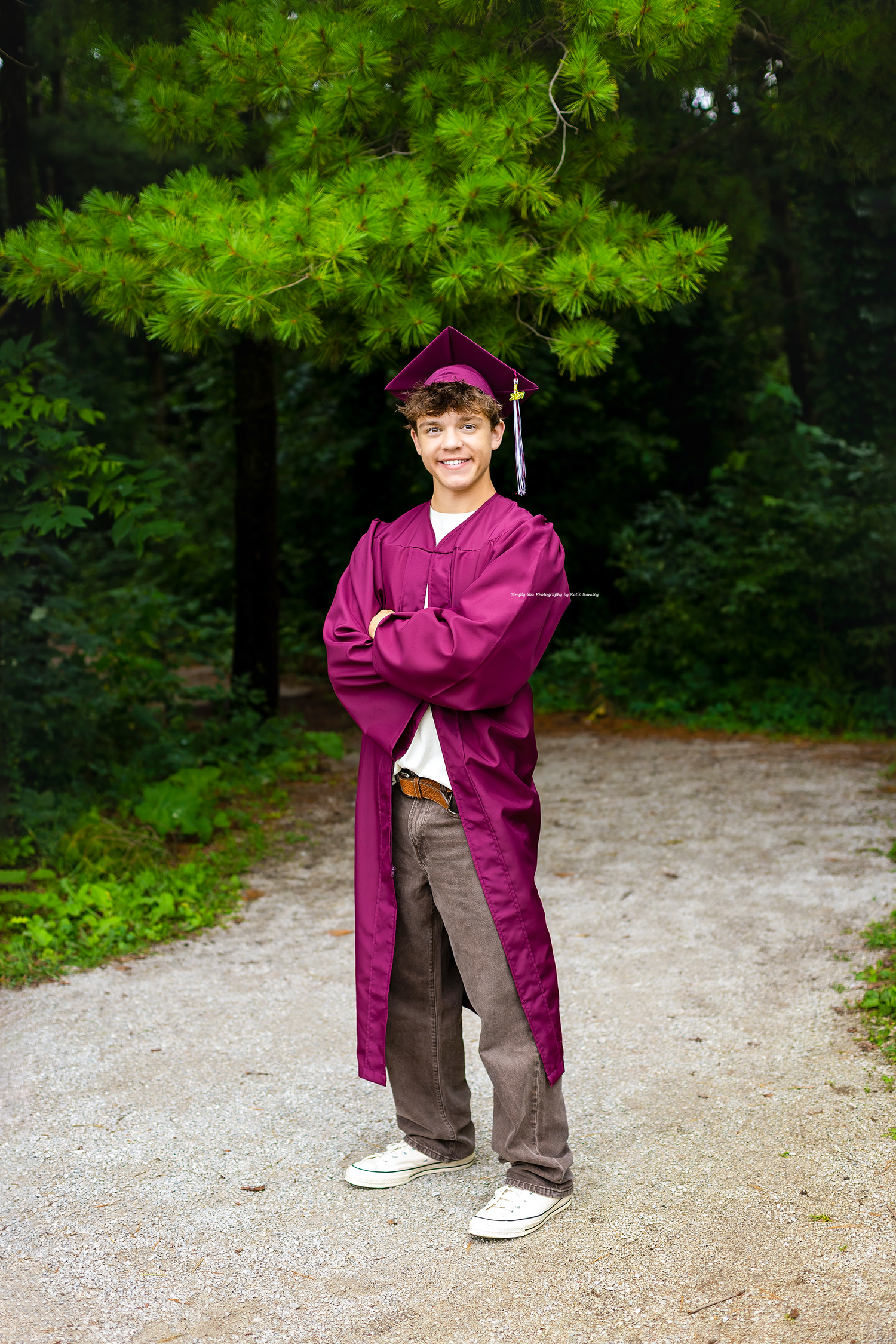 Smiling teen in maroon graduation cap and gown standing with arms crossed on a gravel path in a forest.