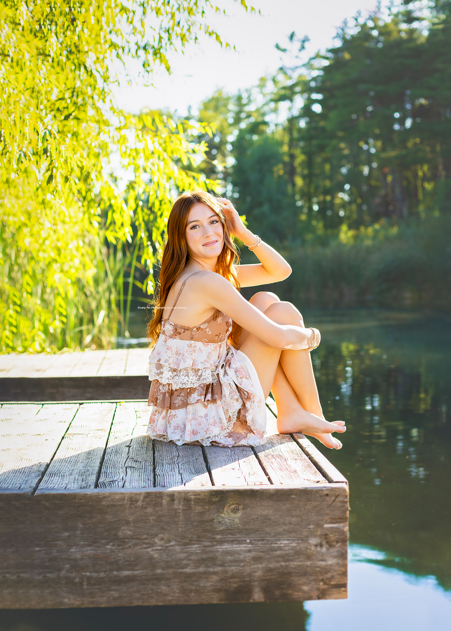 Young woman in floral dress sitting on wooden dock by calm lake surrounded by lush green trees in golden sunlight.