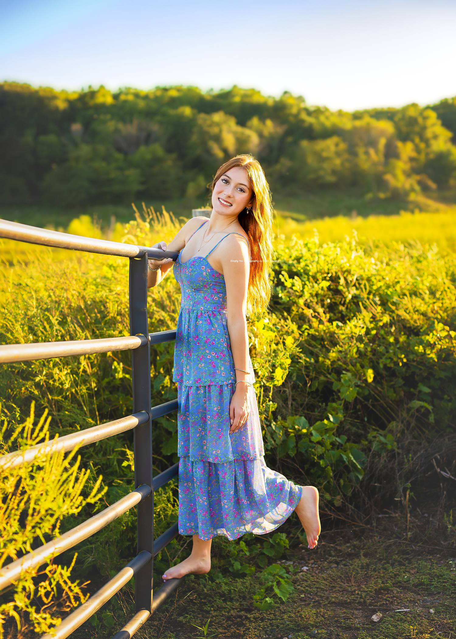Young woman in blue floral midi dress leaning on fence in sunlit green field at golden hour.