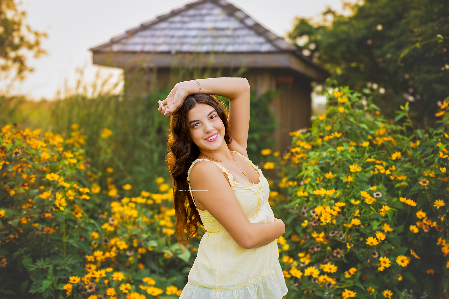 Young woman in yellow dress posing among bright yellow flowers near a gazebo in golden hour sunlight.