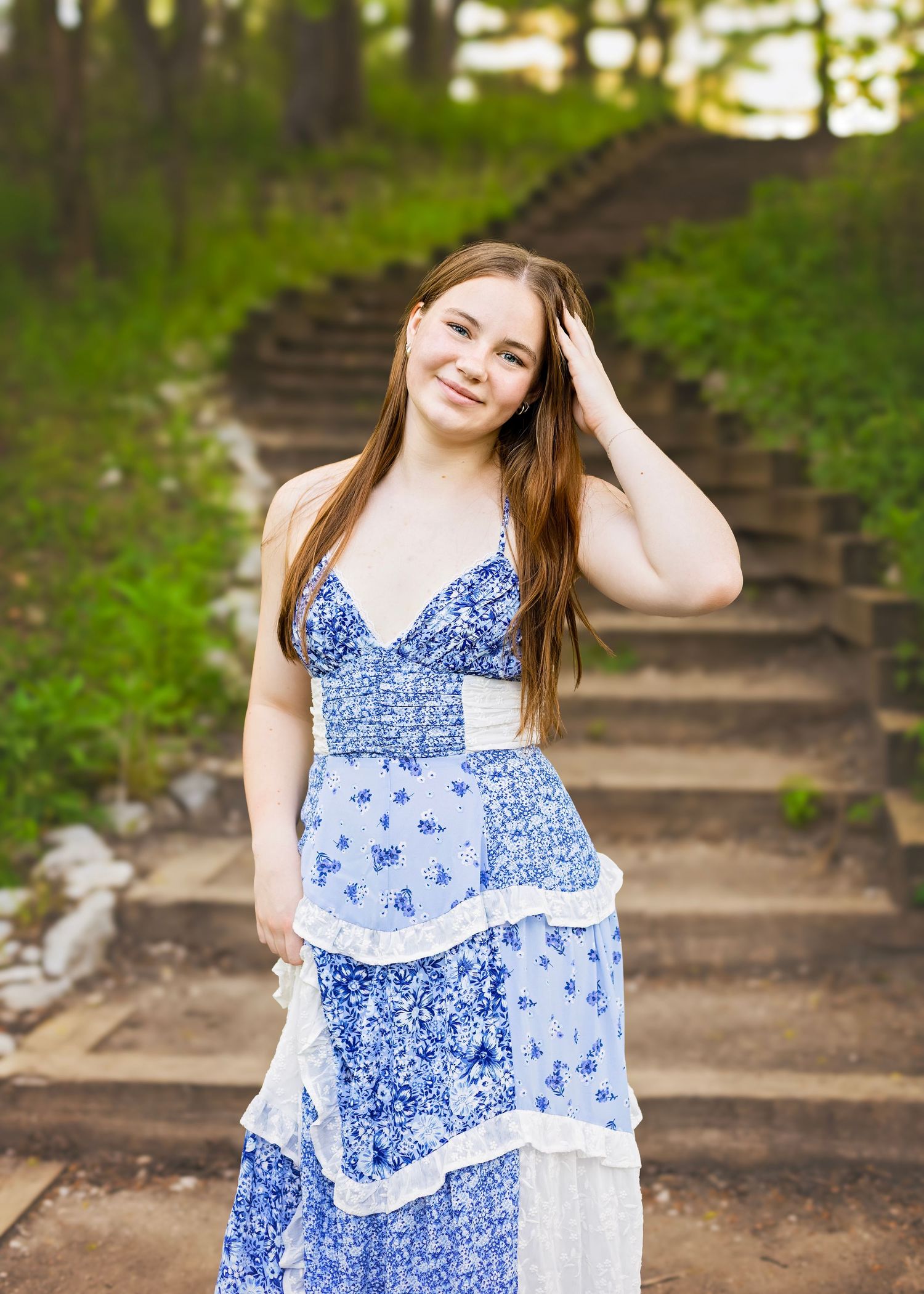 Young woman in blue floral tiered maxi dress poses on stone steps surrounded by lush green trees.