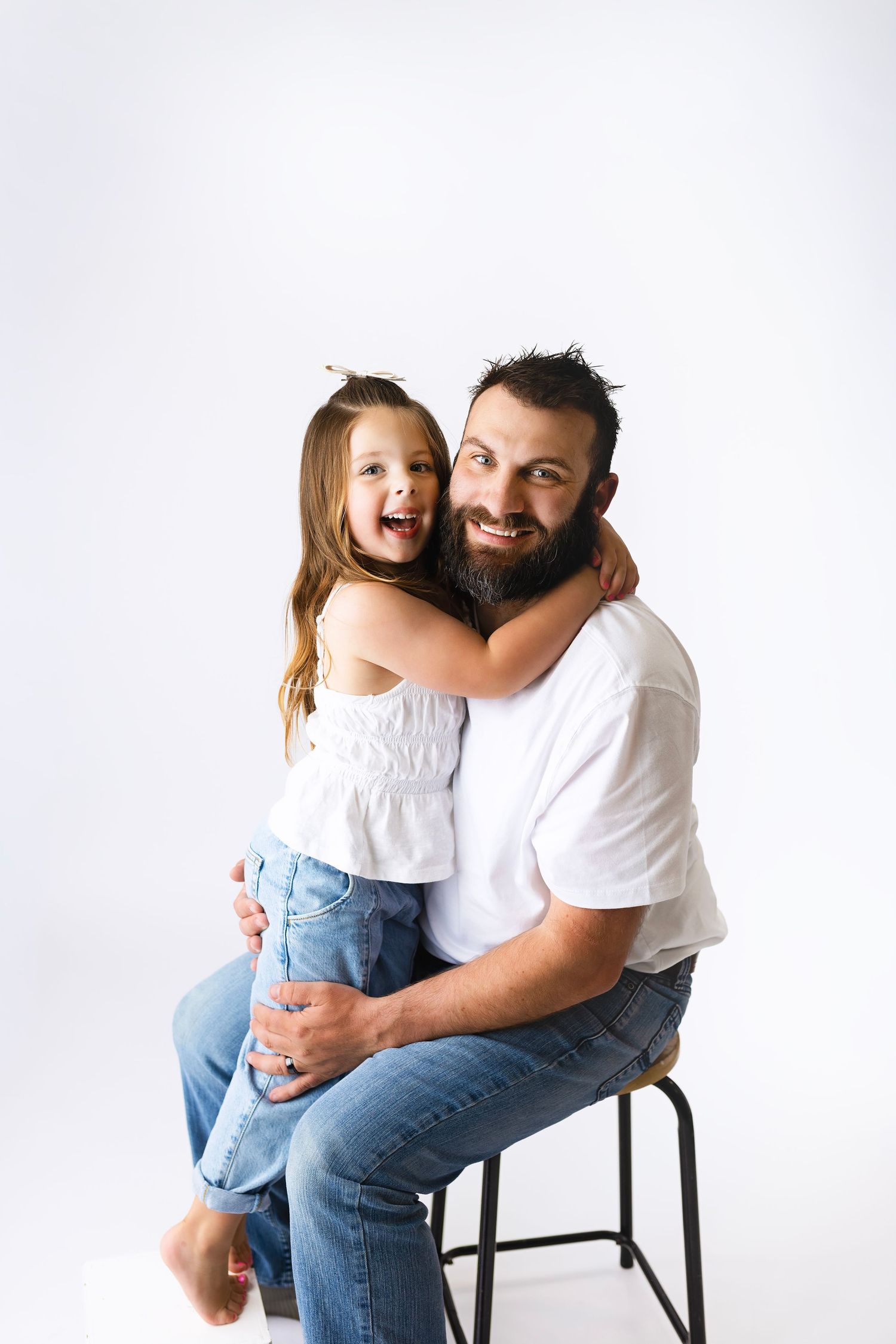A bearded man sitting on a stool hugging a laughing little girl in white top and jeans against white background.