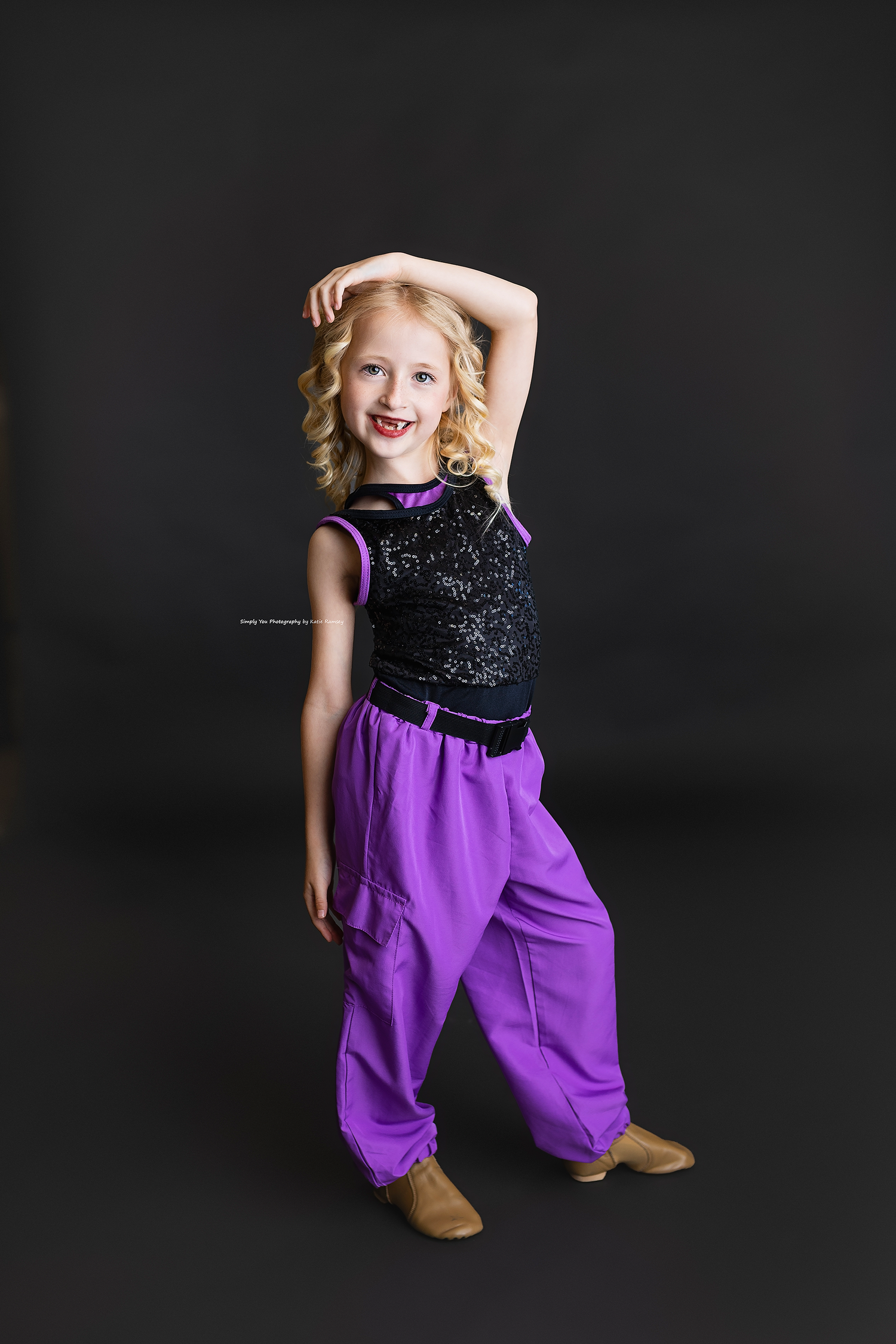 Smiling young blonde girl in purple harem pants and black sequin top poses in a dance stance.