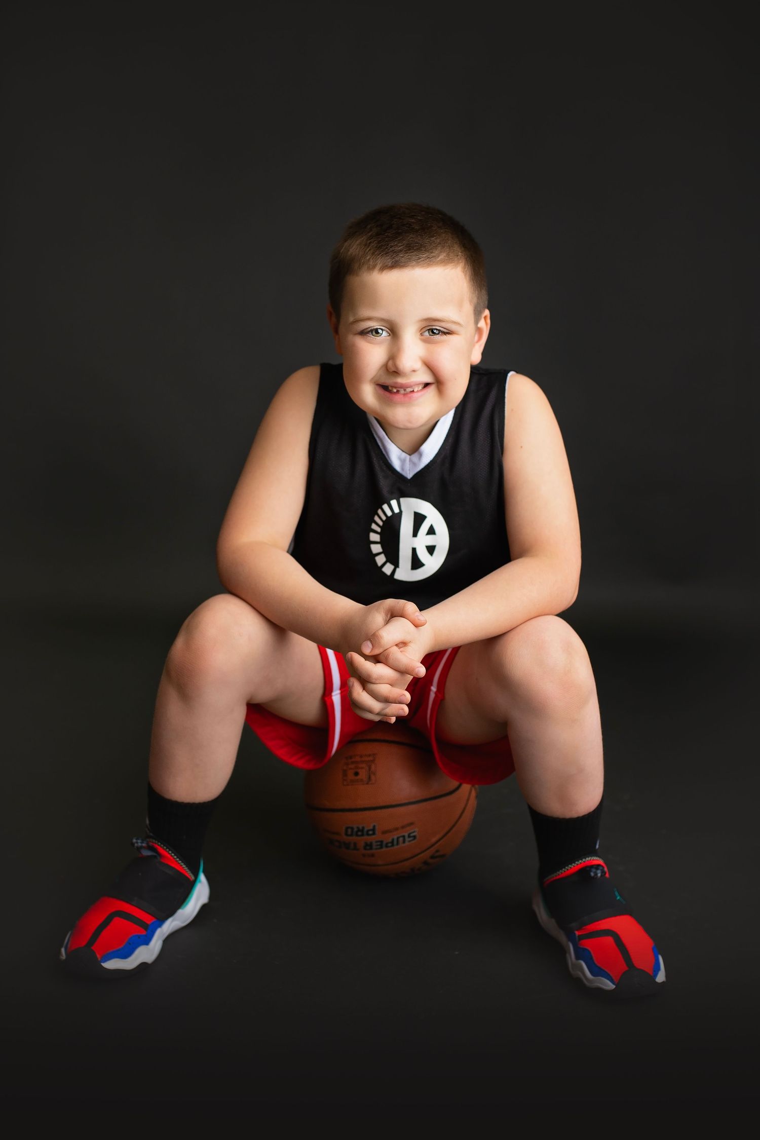 Smiling young boy in Brooklyn Nets basketball uniform sitting on basketball on dark background.