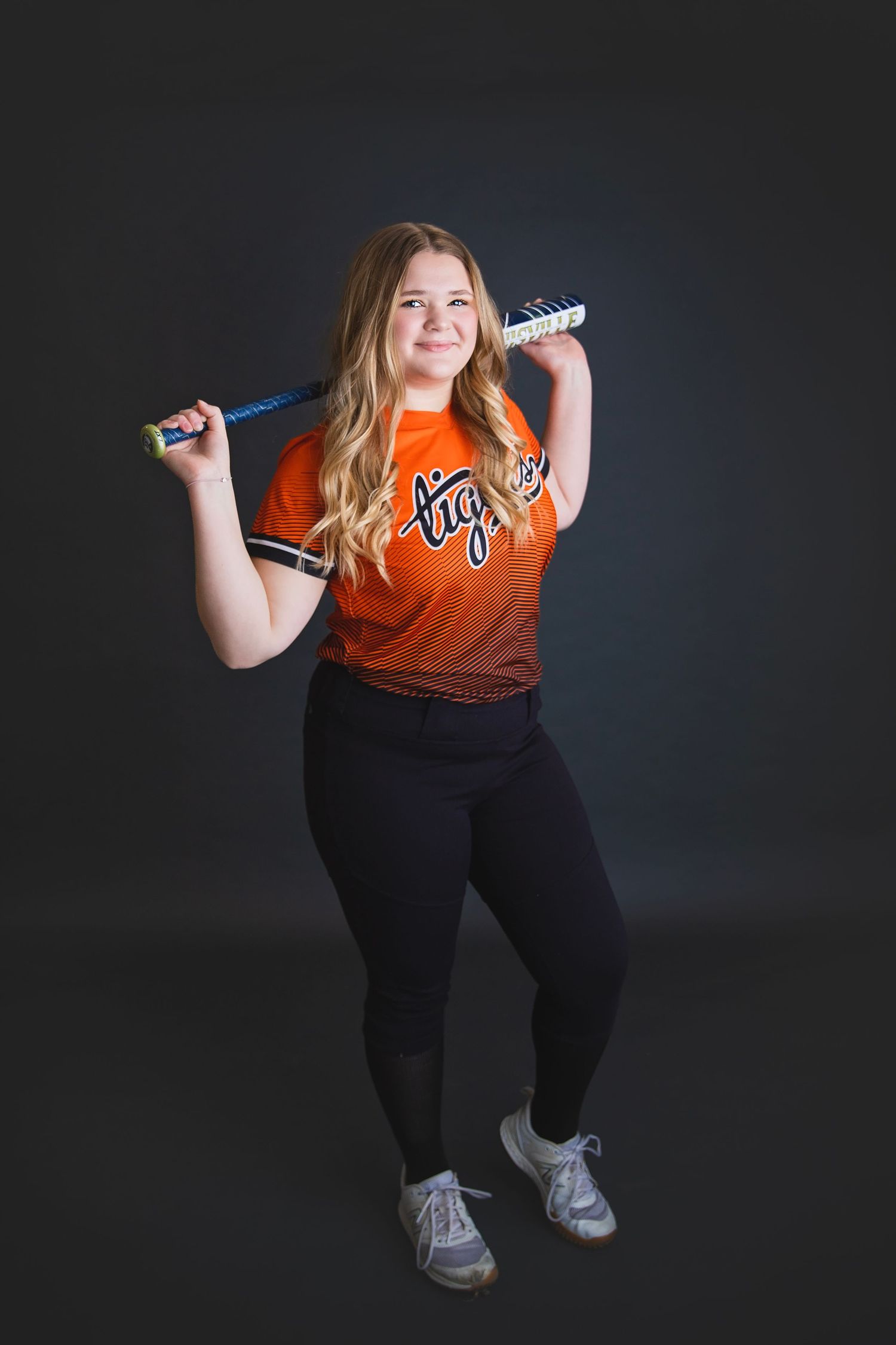 Teen softball player in orange Tigers jersey poses with bat across shoulders in studio portrait.