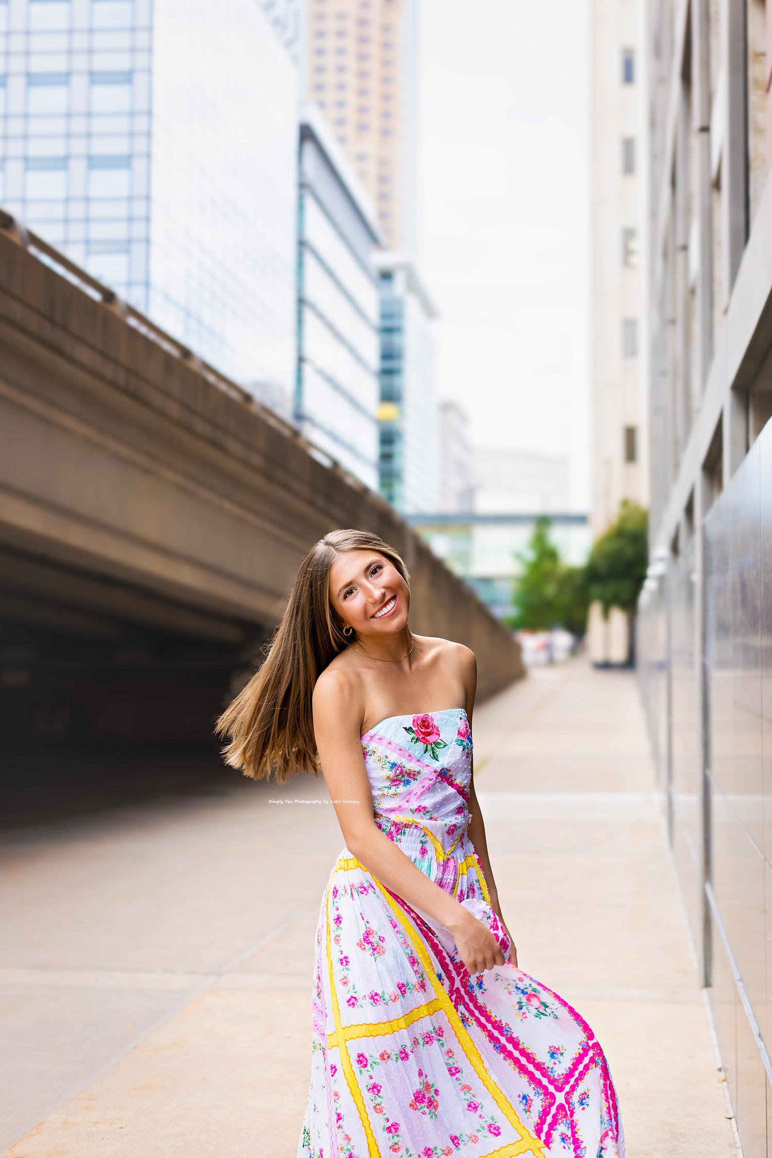 Young woman in floral strapless maxi dress smiling outdoors in urban city setting with modern buildings.