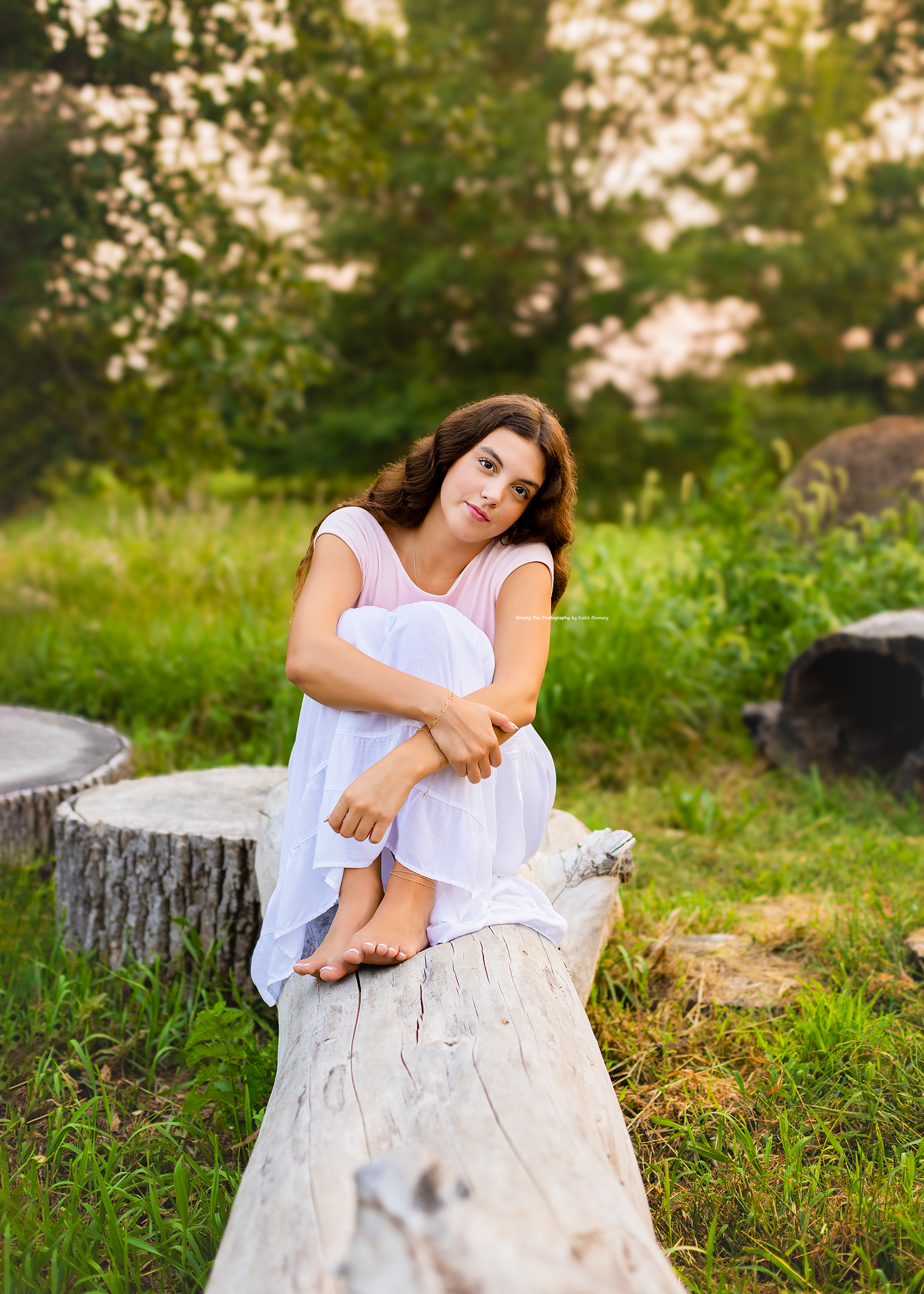 Young woman in white dress sitting on a log in a lush green outdoor setting during golden hour.