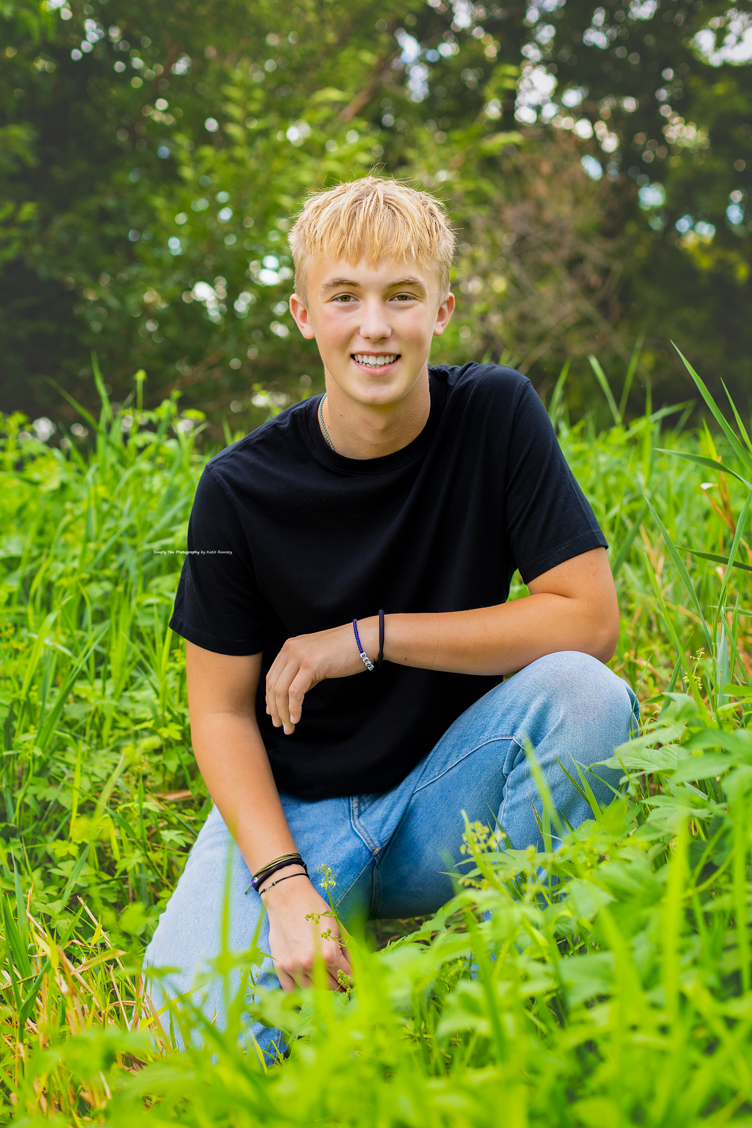 A smiling blonde teenage boy sits in lush green grass wearing a black t-shirt and jeans outdoors.