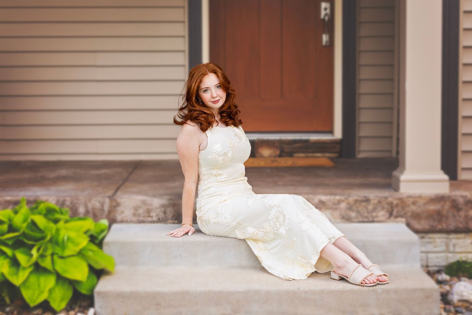 Young redhead woman in elegant white dress sitting on front porch steps for senior portrait.