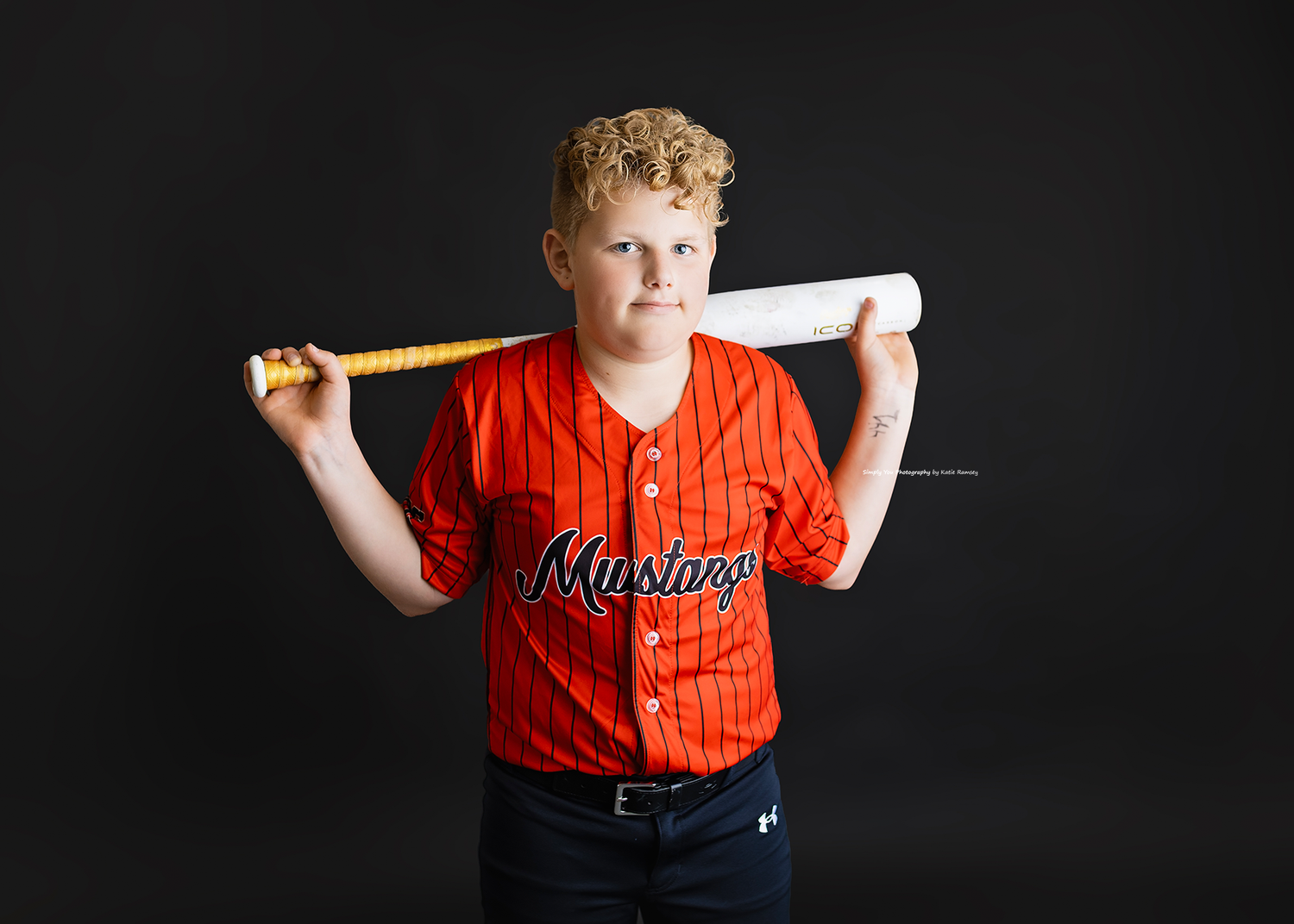 Young boy in red Mustangs baseball jersey holding bat over shoulders.