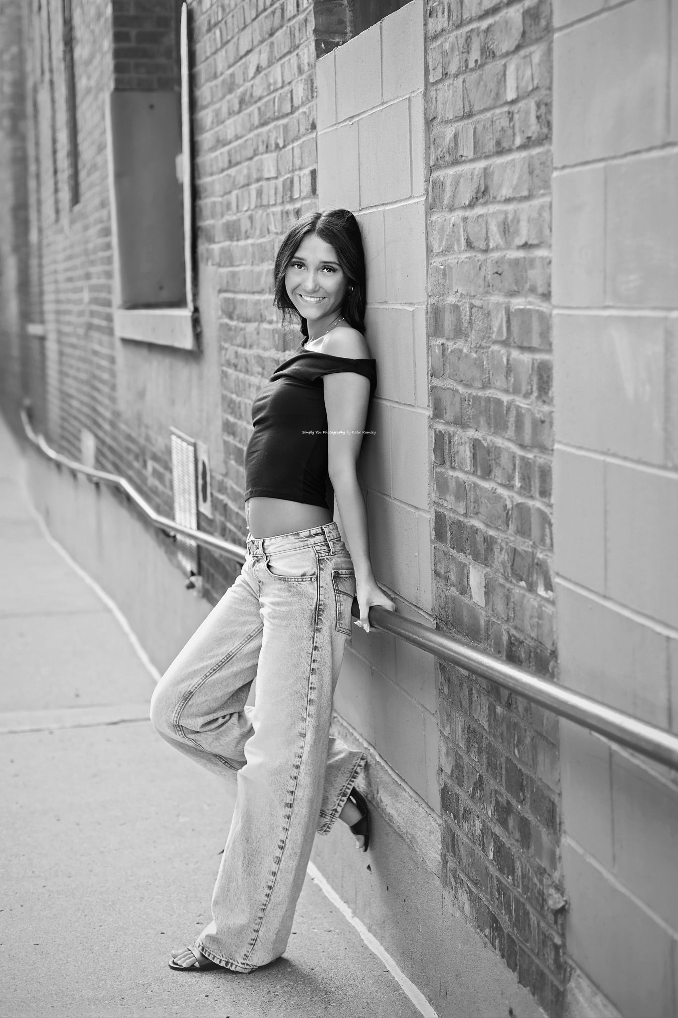 Young woman in black crop top and wide-leg jeans leaning against brick wall in urban alley, black and white photo.