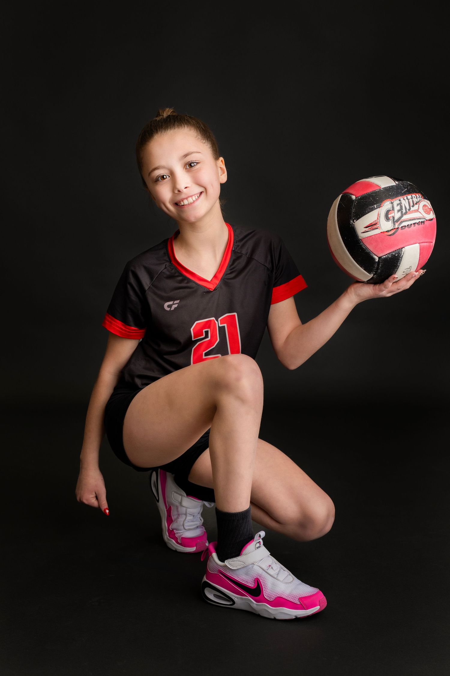Smiling girl in number 21 black volleyball uniform kneeling and holding Central volleyball in studio.