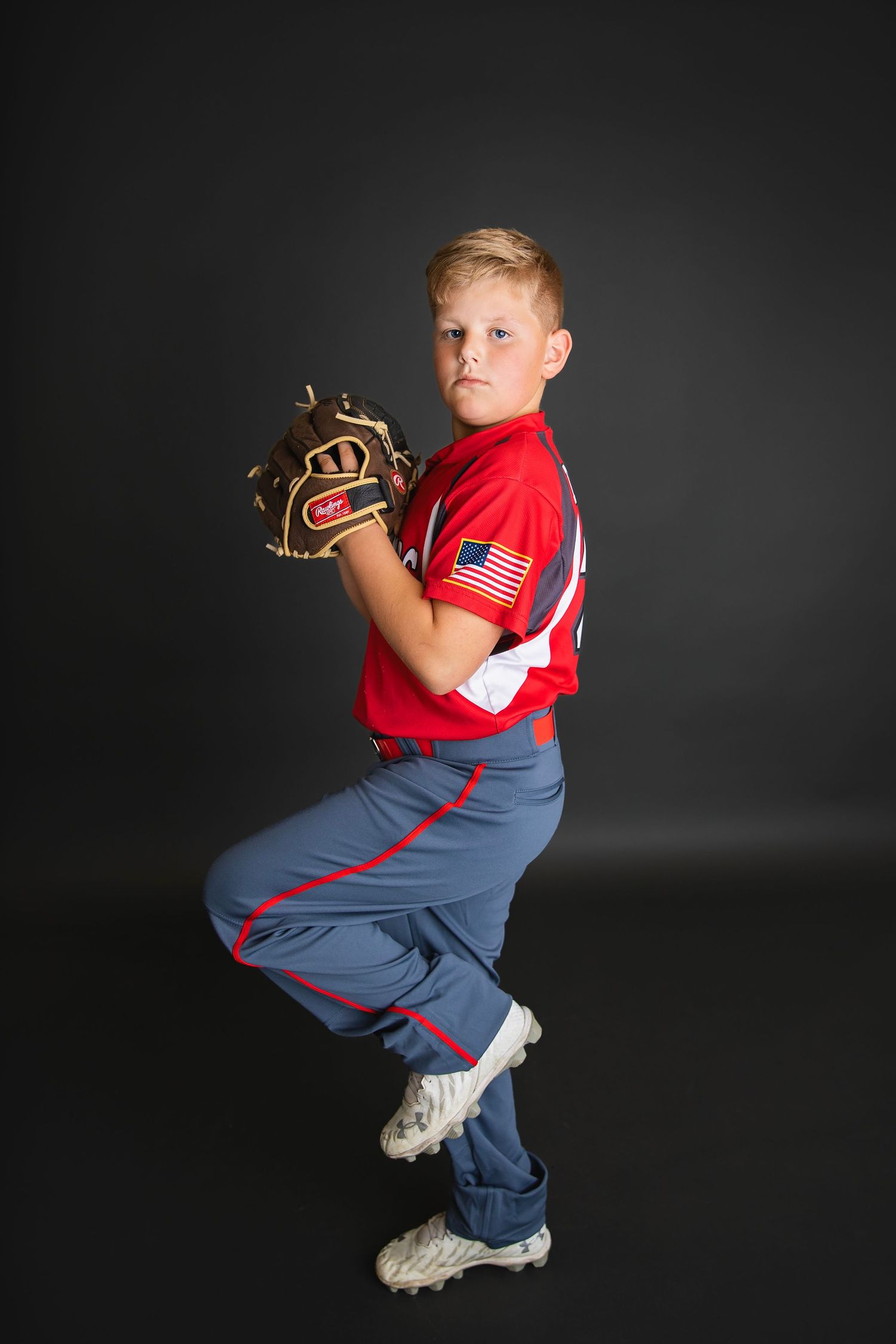 Young baseball player in red Mustangs jersey with American flag patch poses in full pitching windup stance.