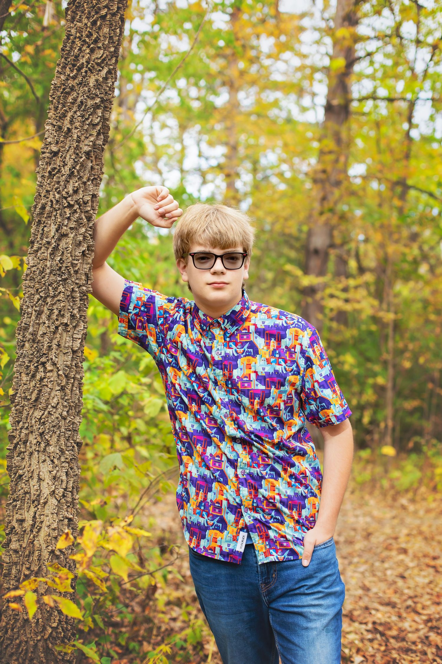 Teenage boy in colorful patterned shirt leaning against a tree in autumn forest.