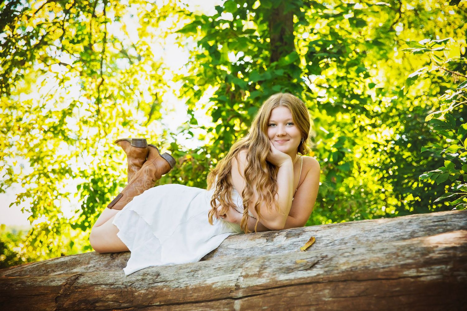 Smiling teen girl in white dress lies on a log surrounded by lush green trees.