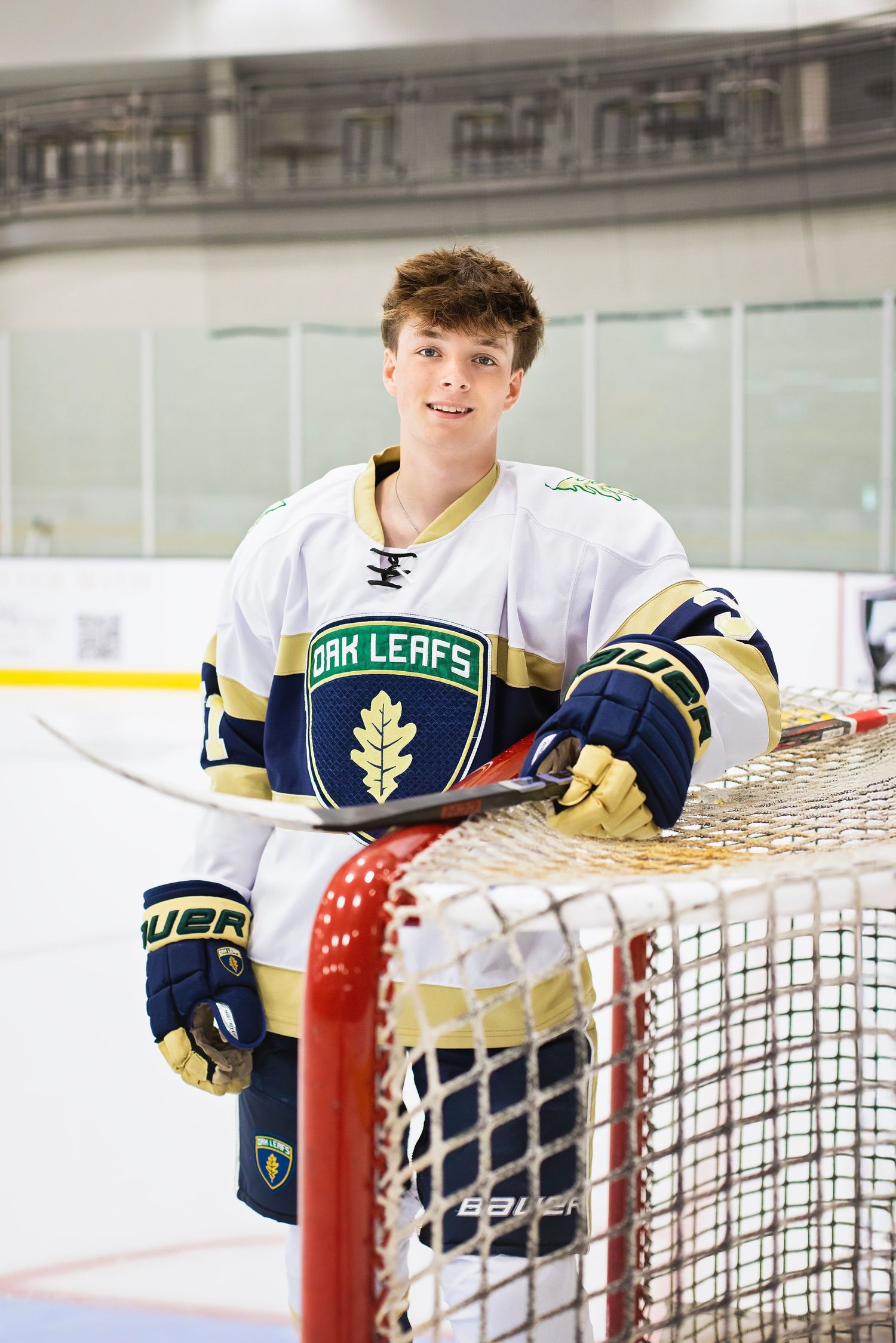 Teen hockey player in Oak Leafs jersey smiling while leaning on goal net at rink.