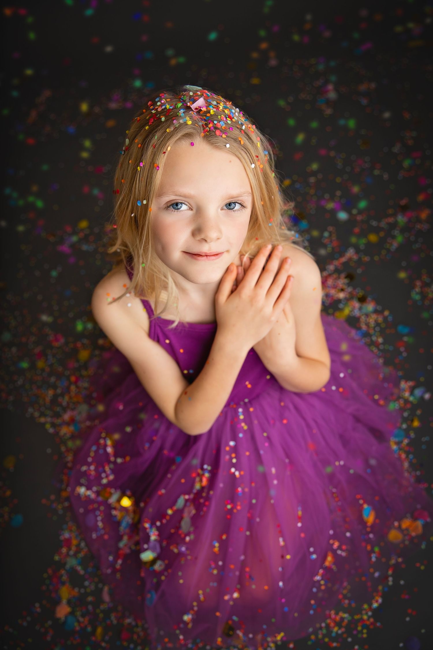 Sweet blonde girl in purple tutu looking up with hands clasped amid colorful confetti bokeh.