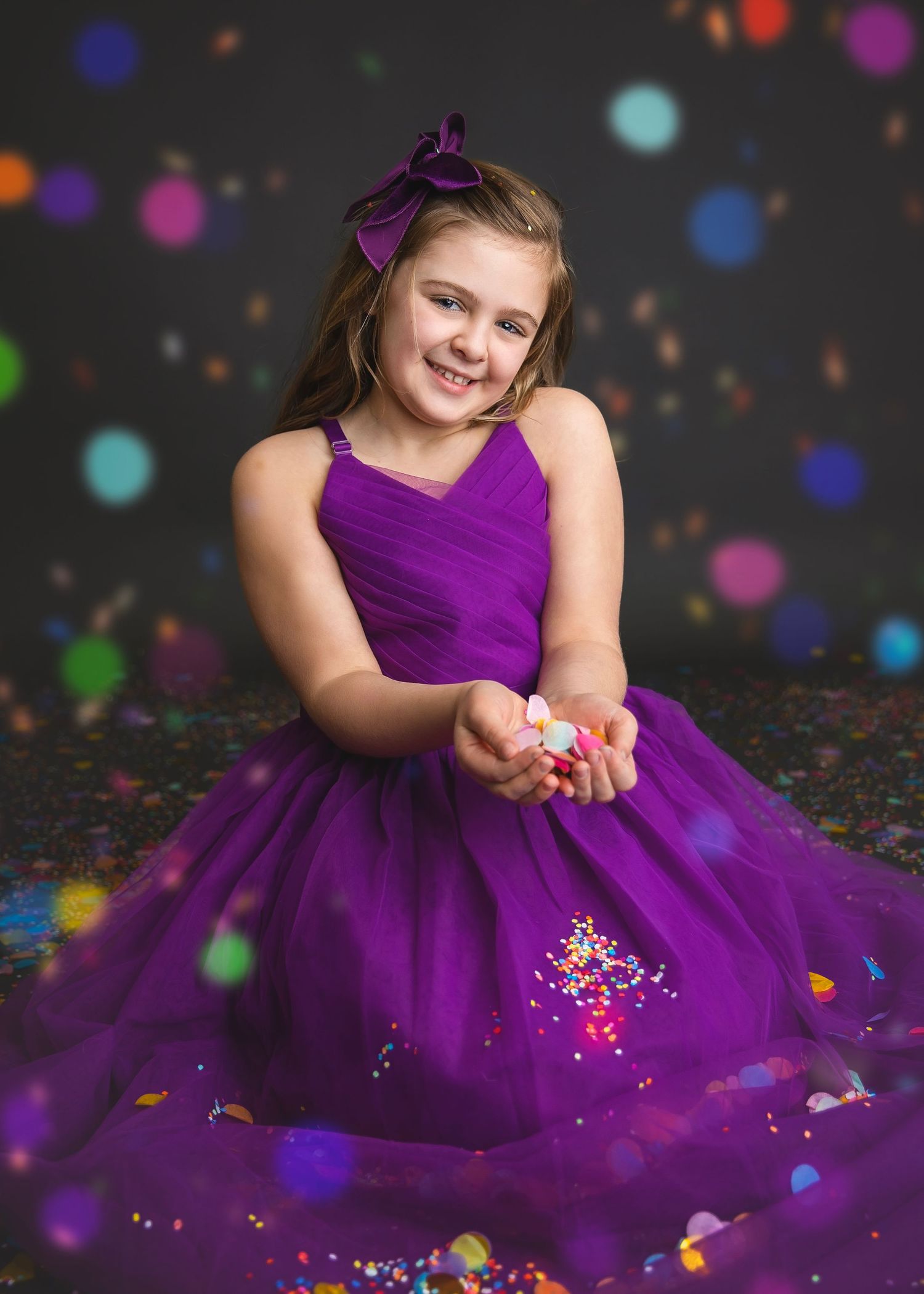 Smiling girl in purple tulle dress sitting on floor, holding confetti in hands during studio shoot.