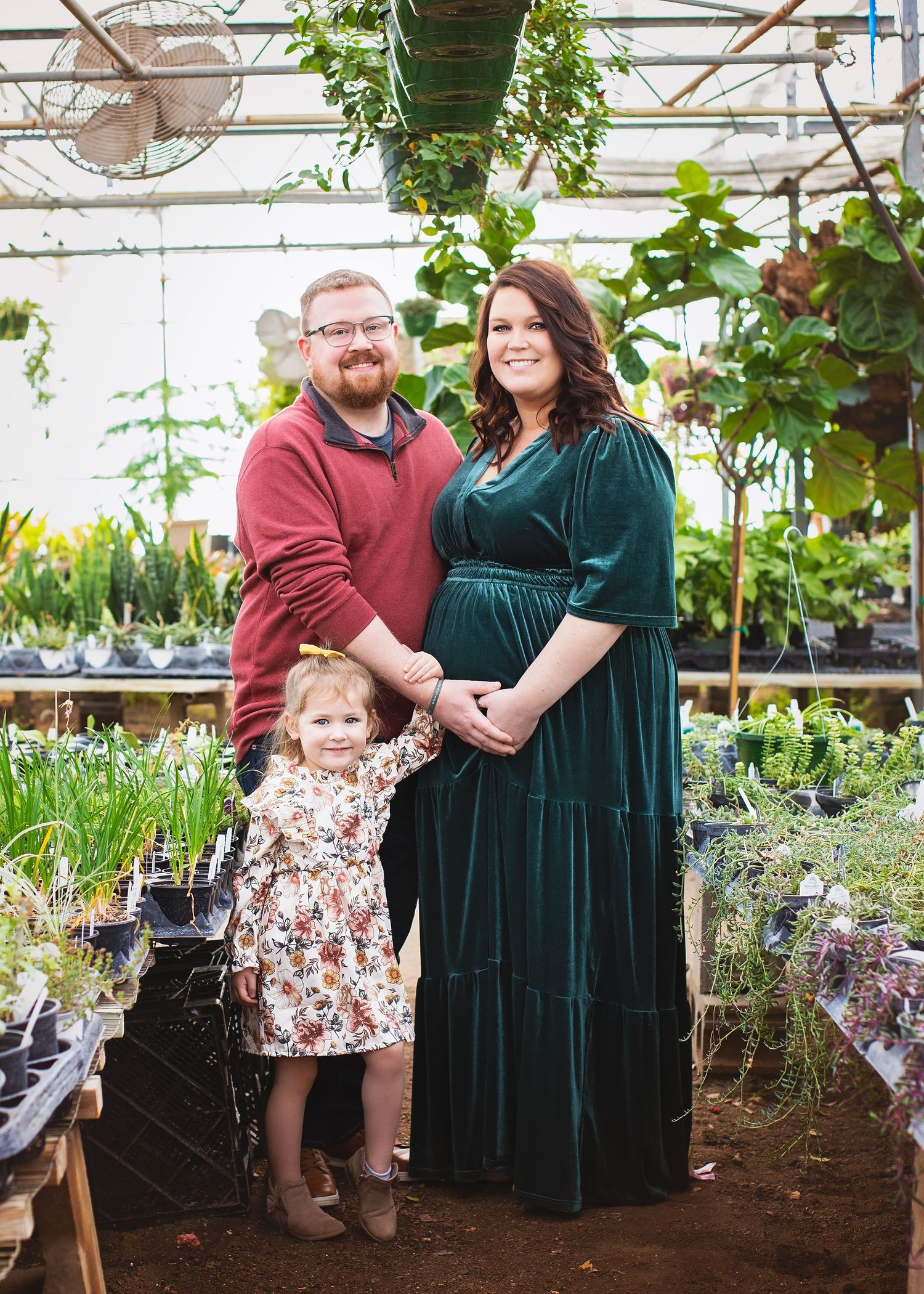 Expecting couple with toddler daughter pose for maternity photos inside a lush plant nursery greenhouse.