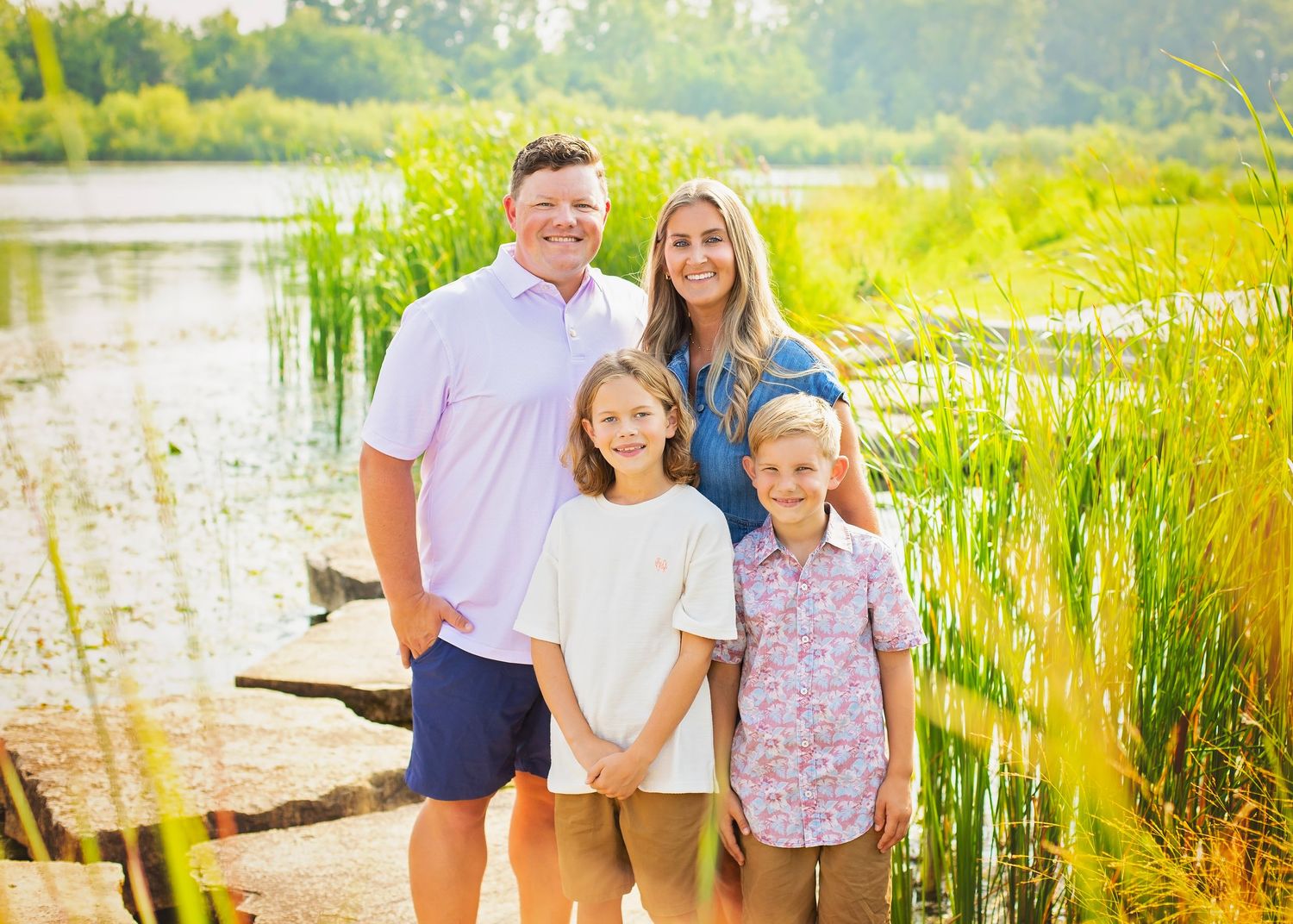 Family of four posing together near a pond surrounded by tall green reeds in golden sunlight.