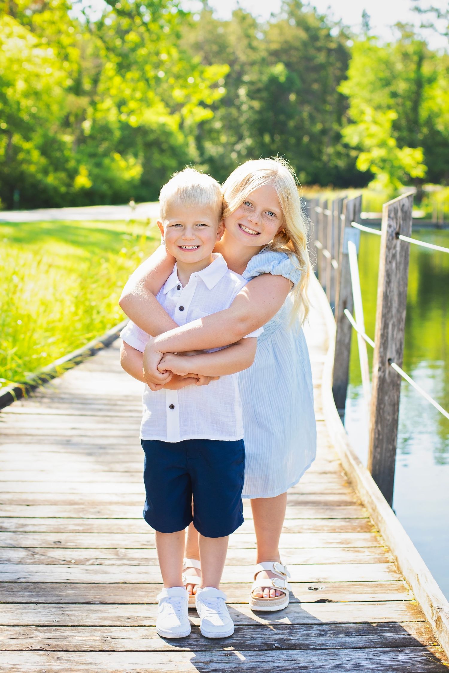 Blonde sister hugs younger brother on a wooden dock surrounded by lush green trees and a pond.