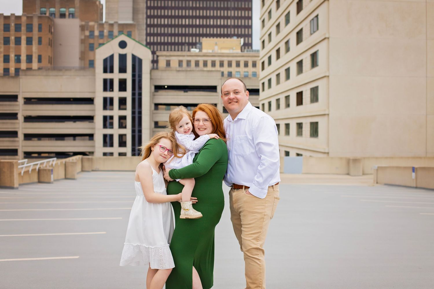 Smiling family of four with pregnant mother in green dress hugging on urban rooftop location.
