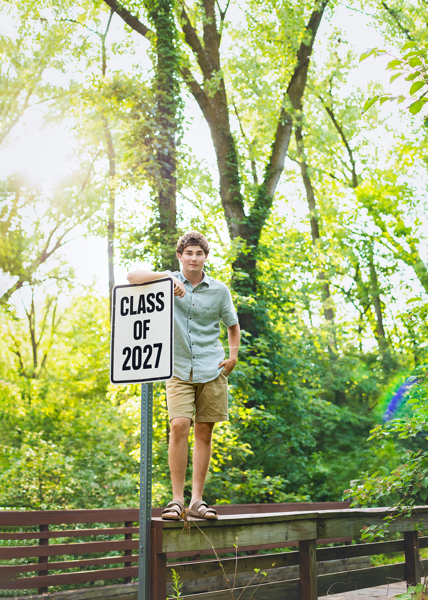 A Class of 2027 student poses proudly on a fence beside a street sign surrounded by lush green trees.