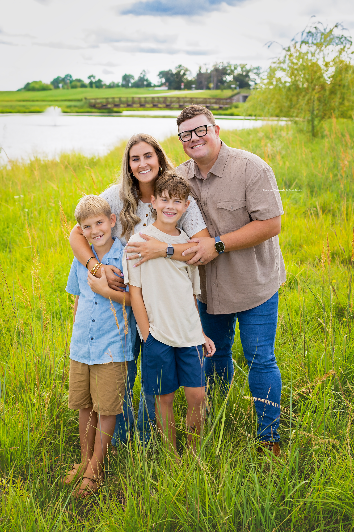 Happy family of four posing outdoors in lush green grass near a pond on a sunny day.
