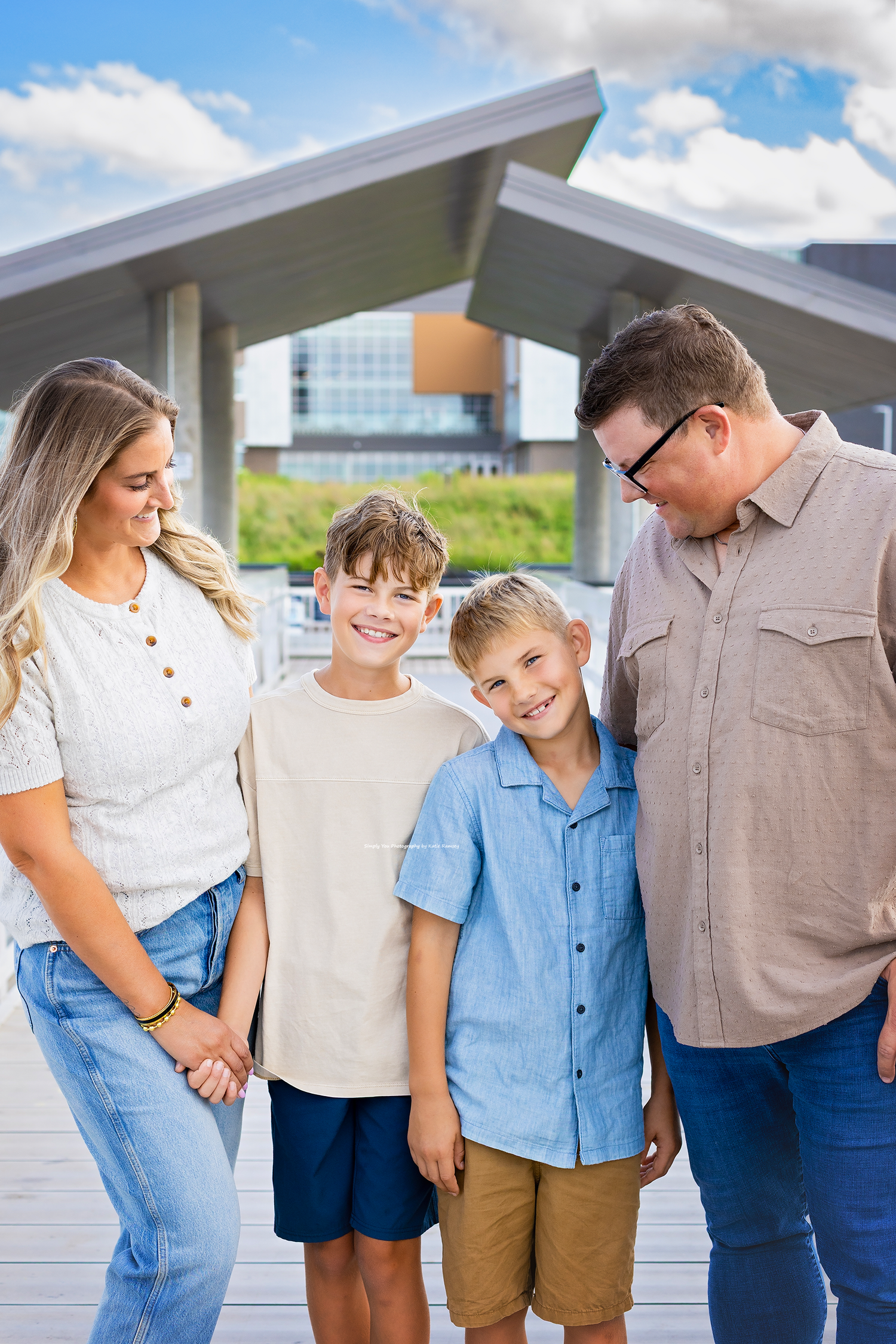 A happy family of four poses outdoors near a modern pavilion under a blue sky with clouds.
