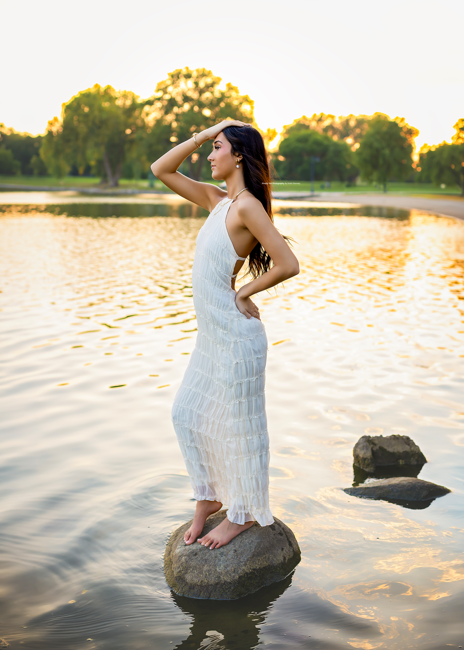 Woman in white dress standing on rock in lake at golden hour sunset with trees in background.