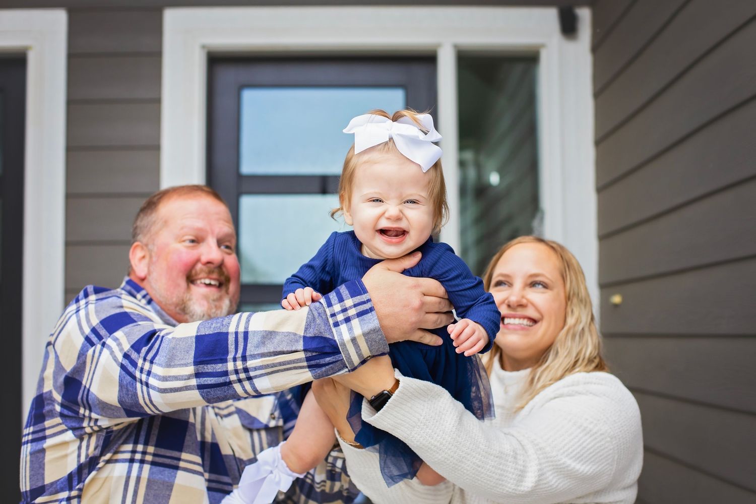 Joyful parents lifting their laughing baby girl in a navy dress outside their home.