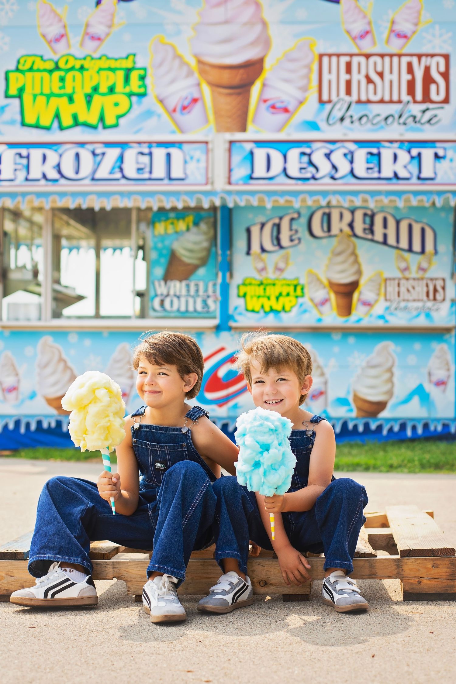 Two boys in matching denim overalls holding colorful cotton candy at a frozen dessert fair stand.