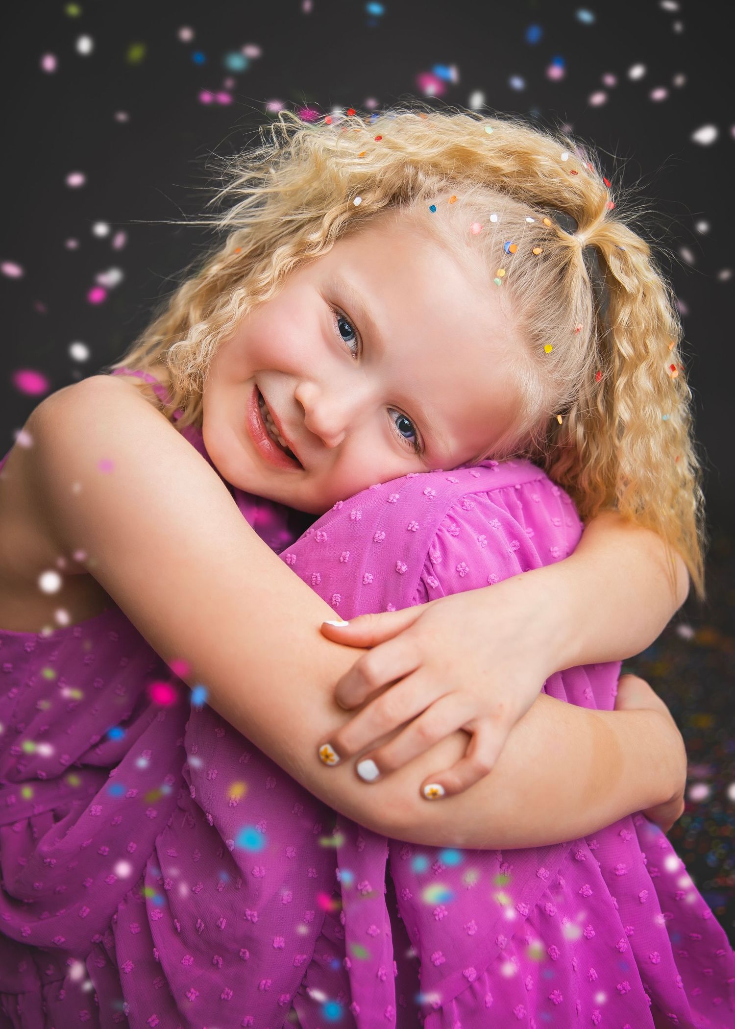 Smiling blonde girl in pink dress hugging herself with colorful confetti on dark background.