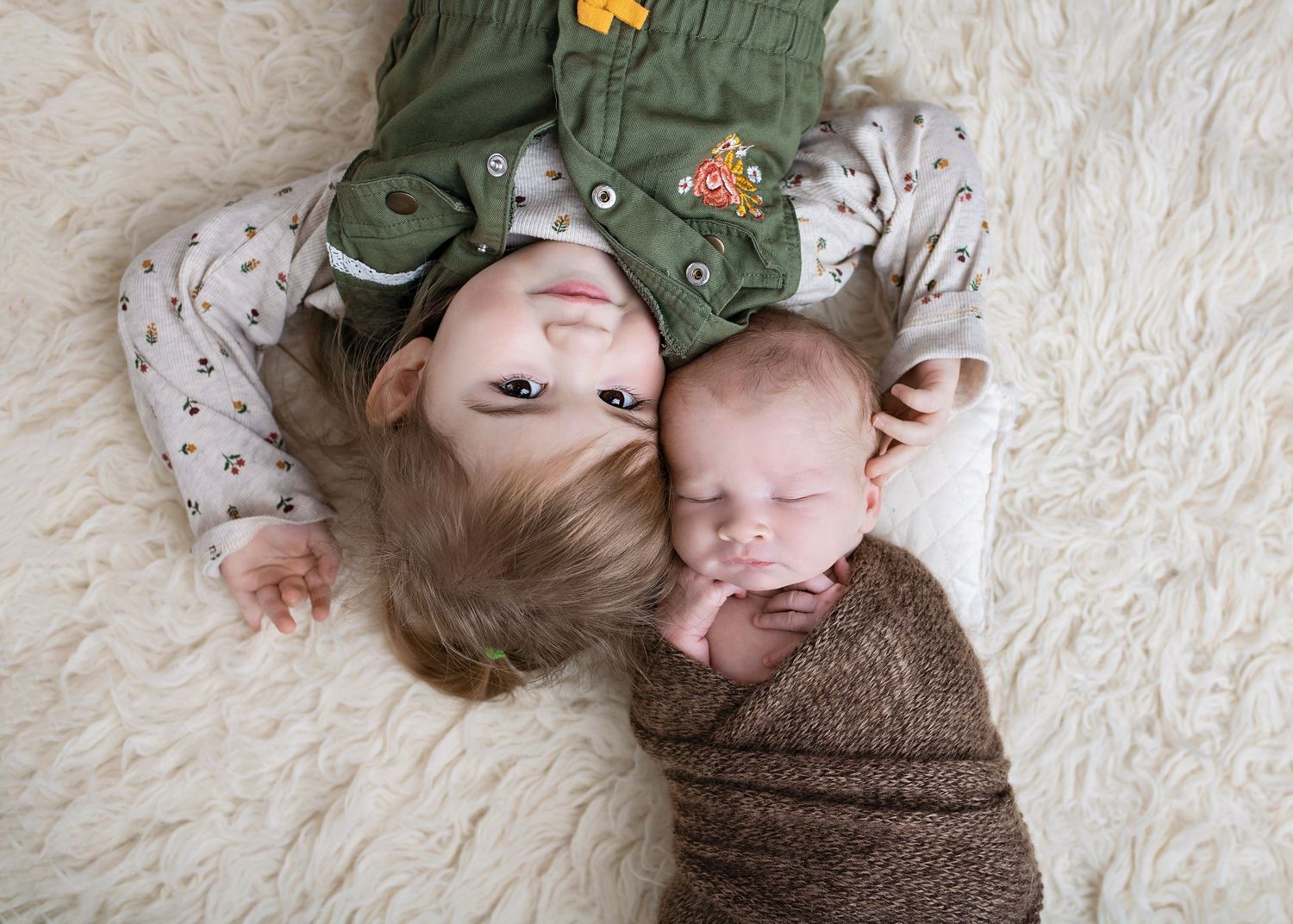 Toddler sibling lying on a white fur rug smiling next to a sleeping newborn wrapped in brown knit.