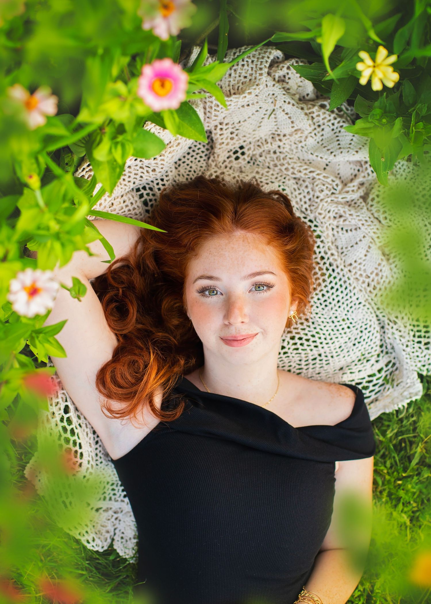 Redhead girl with freckles lying in green grass surrounded by colorful flowers.