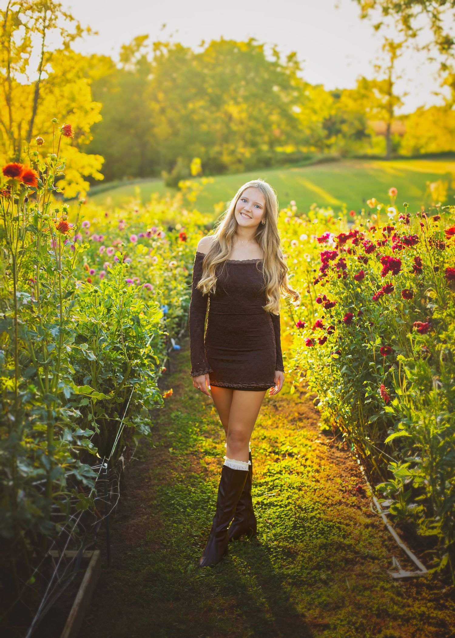 Blonde teen in black mini dress and boots walks through sunlit flower garden rows at golden hour.