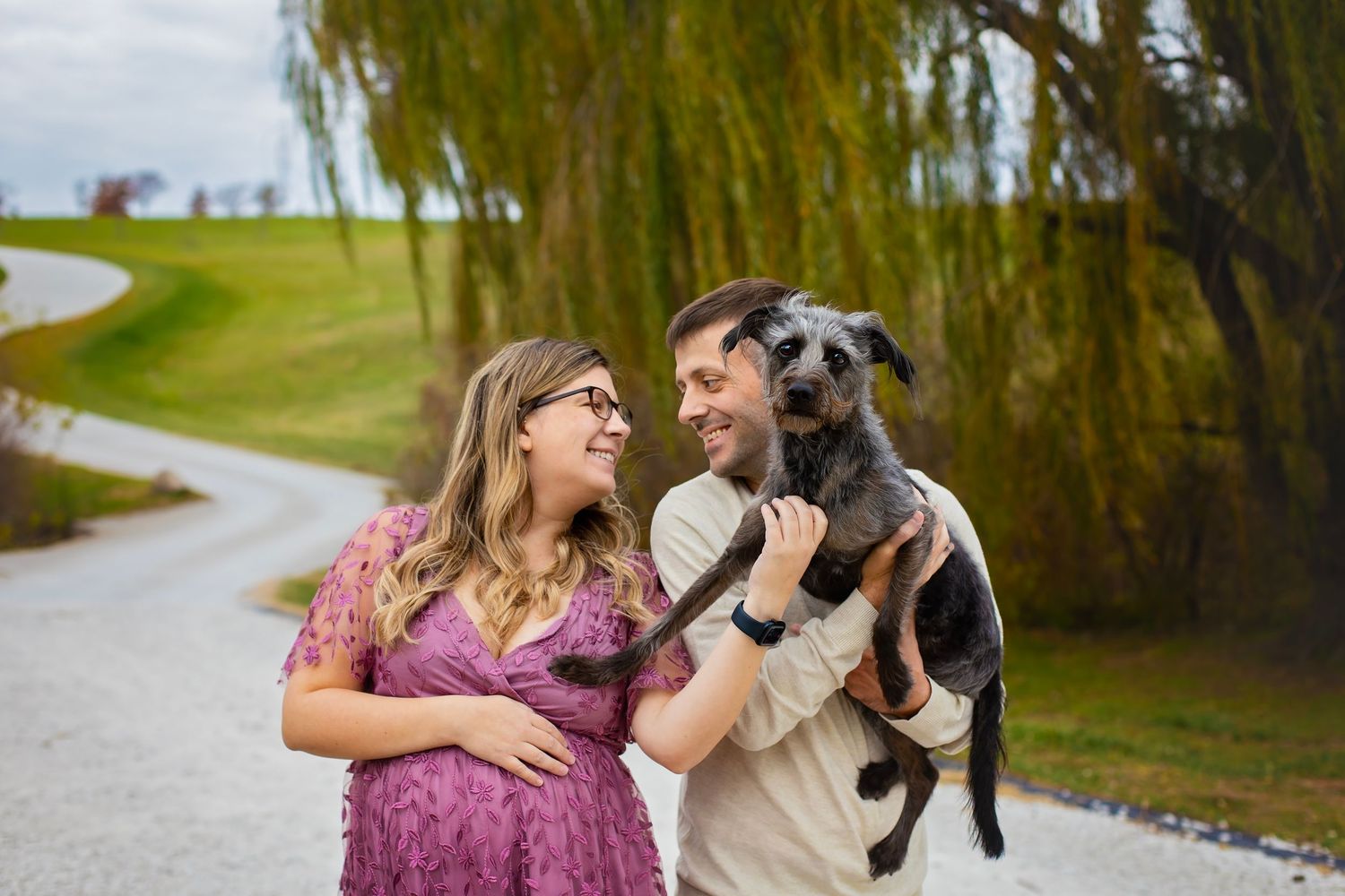 Smiling pregnant couple share a joyful moment with their black dog on a scenic gravel path.