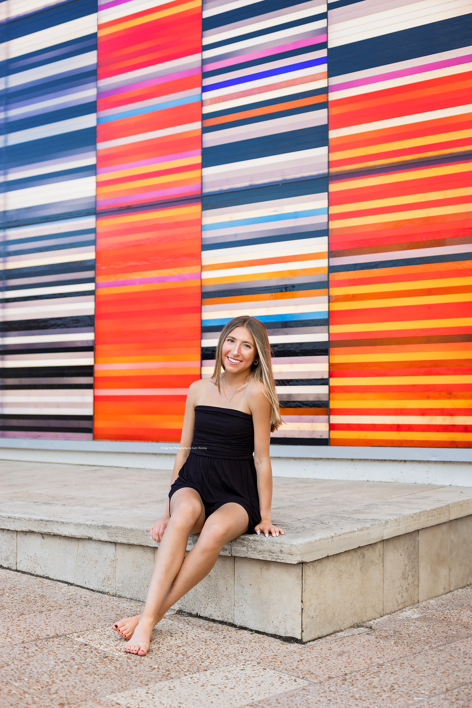 Young woman in black dress sitting barefoot on concrete ledge in front of colorful striped mural wall.