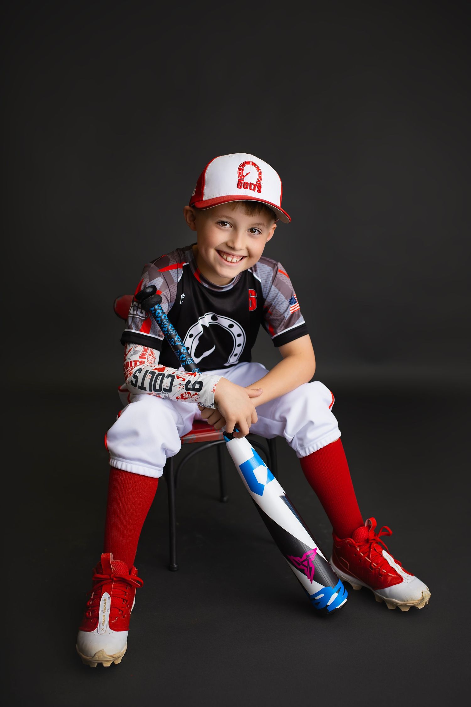 Young baseball player in Colts uniform sitting with bat and smiling on dark background.