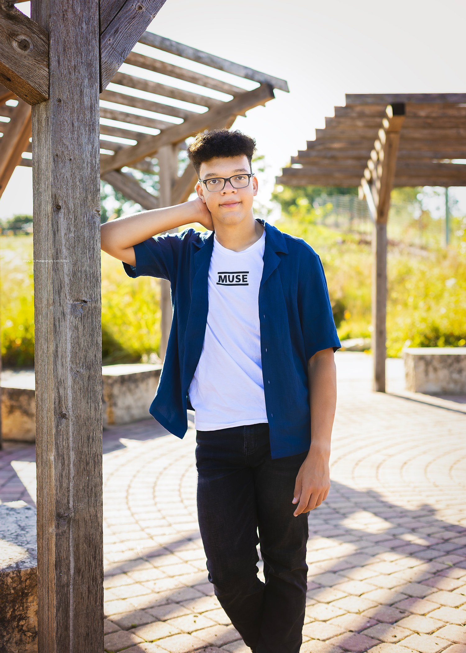 Young man in blue shirt and Muse t-shirt poses outdoors near wooden pergola on brick pathway.