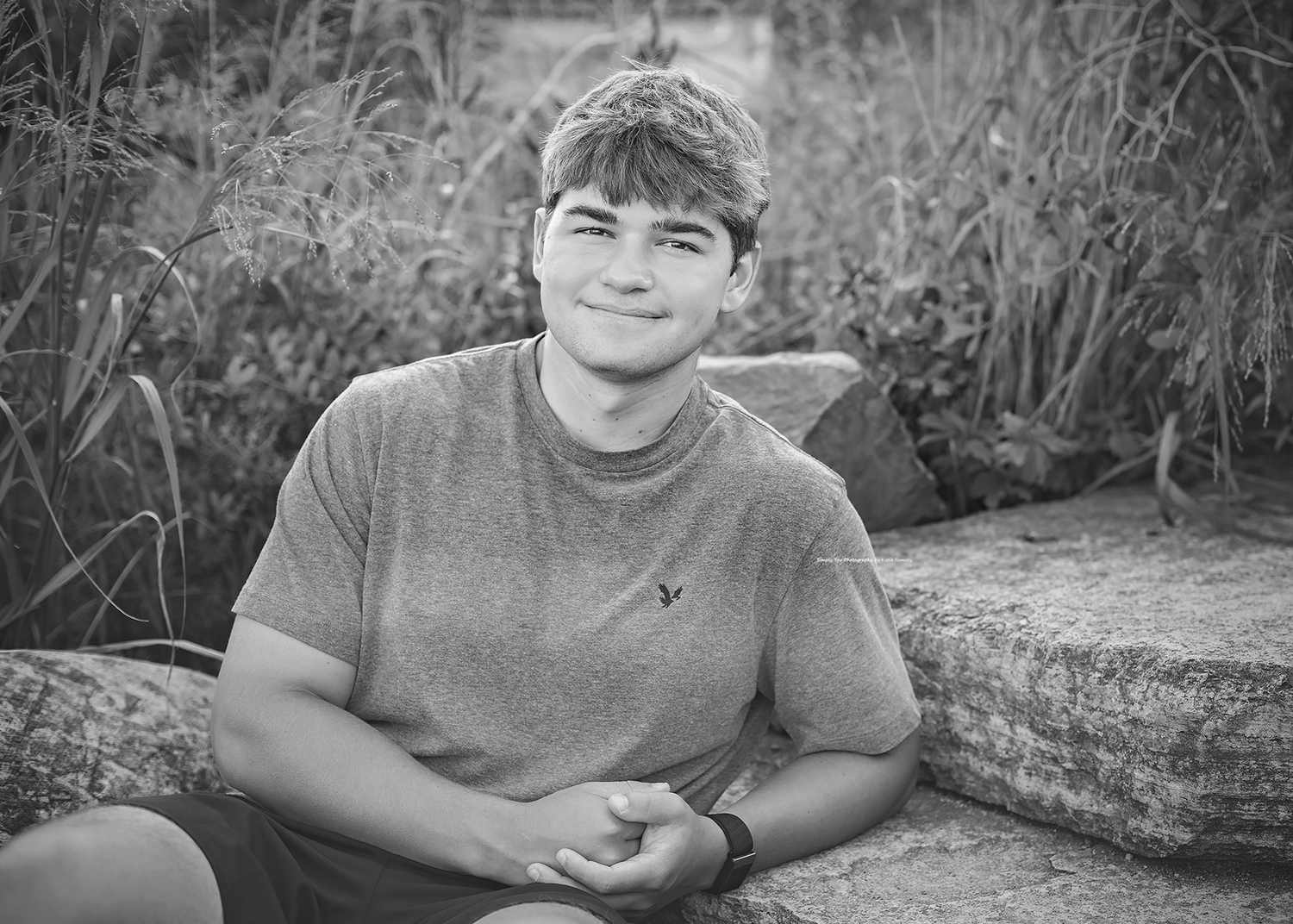 Black and white portrait of a smiling teenage boy sitting on stone steps outdoors among tall grasses.