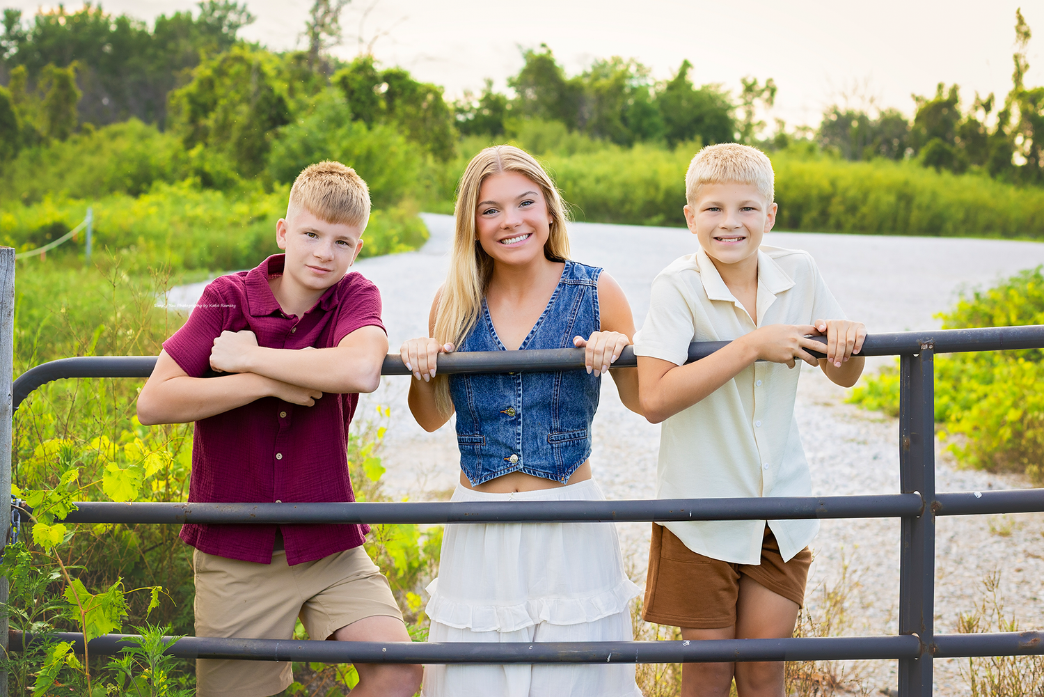 Three smiling kids pose against a metal gate surrounded by lush greenery on a sunny day.